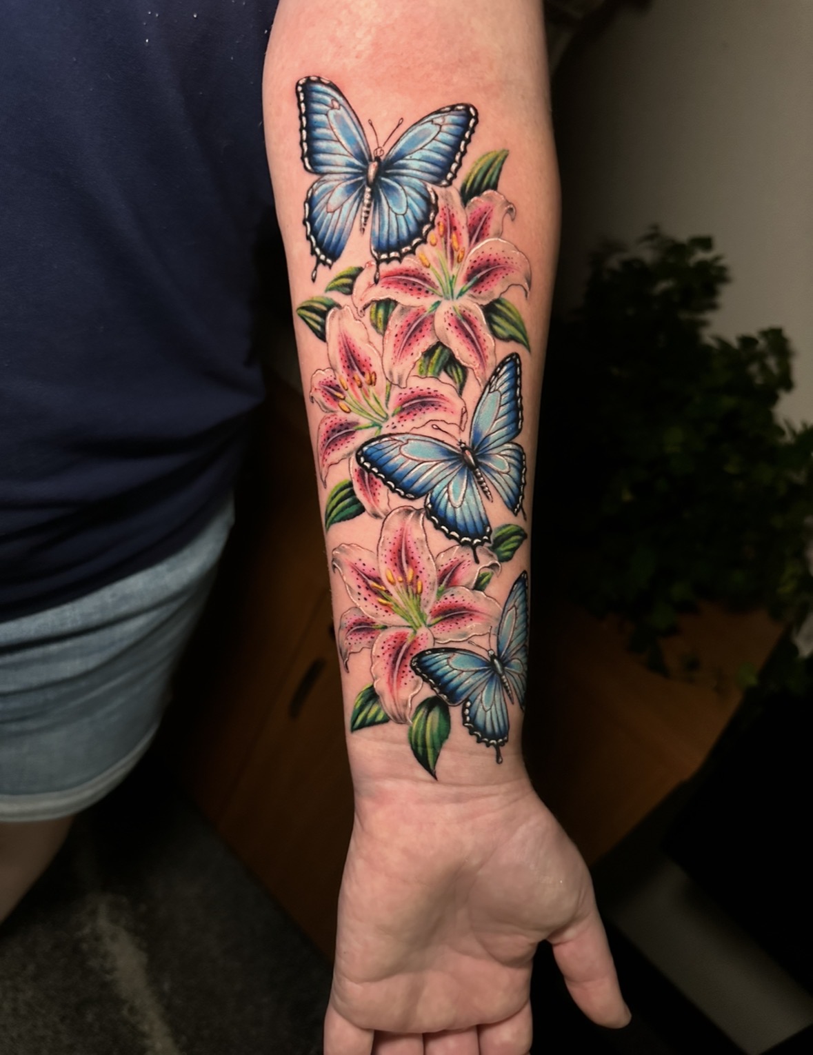 Forearm tattoo with three blue butterflies and pink lilies, detailed and colorful, against a dark background.