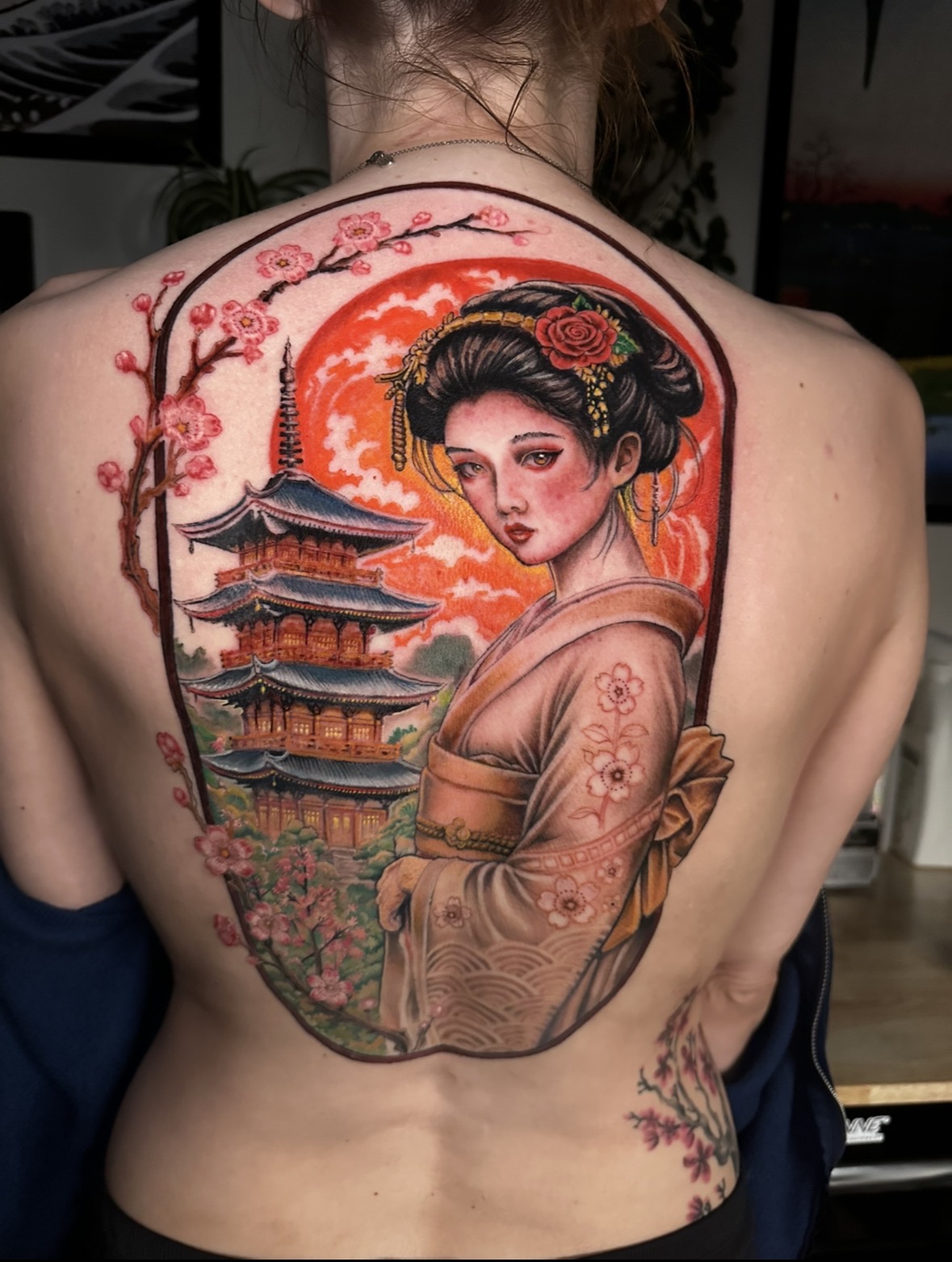 Tattoo of a geisha with a pagoda and cherry blossoms on a person's back, with a sunset background.