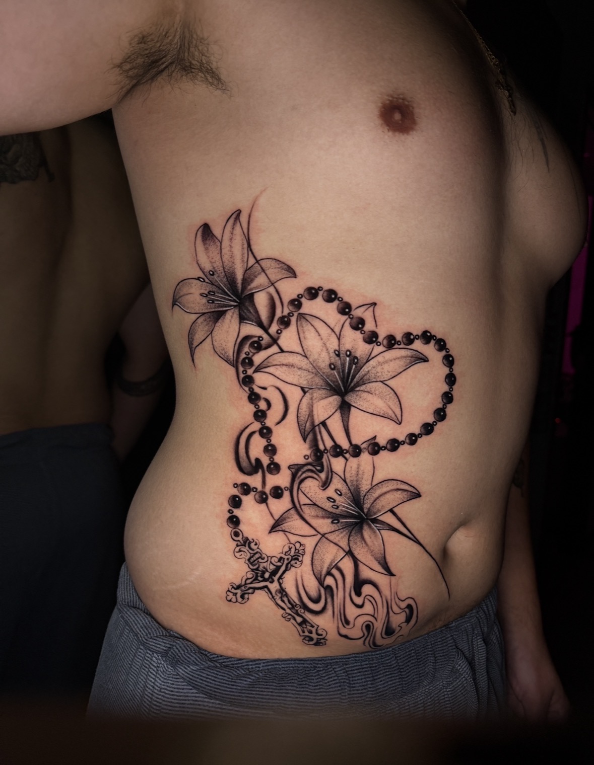 Tattoo of lilies, beads, and a dragon on a person's side and abdomen, with visible skin and chest area.