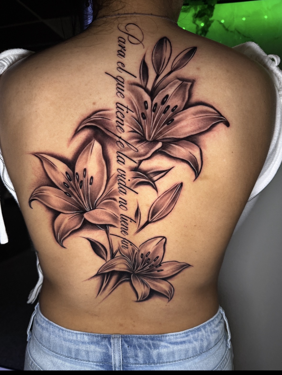 Back tattoo of three large lilies with a quote in cursive script, in black and gray shading.