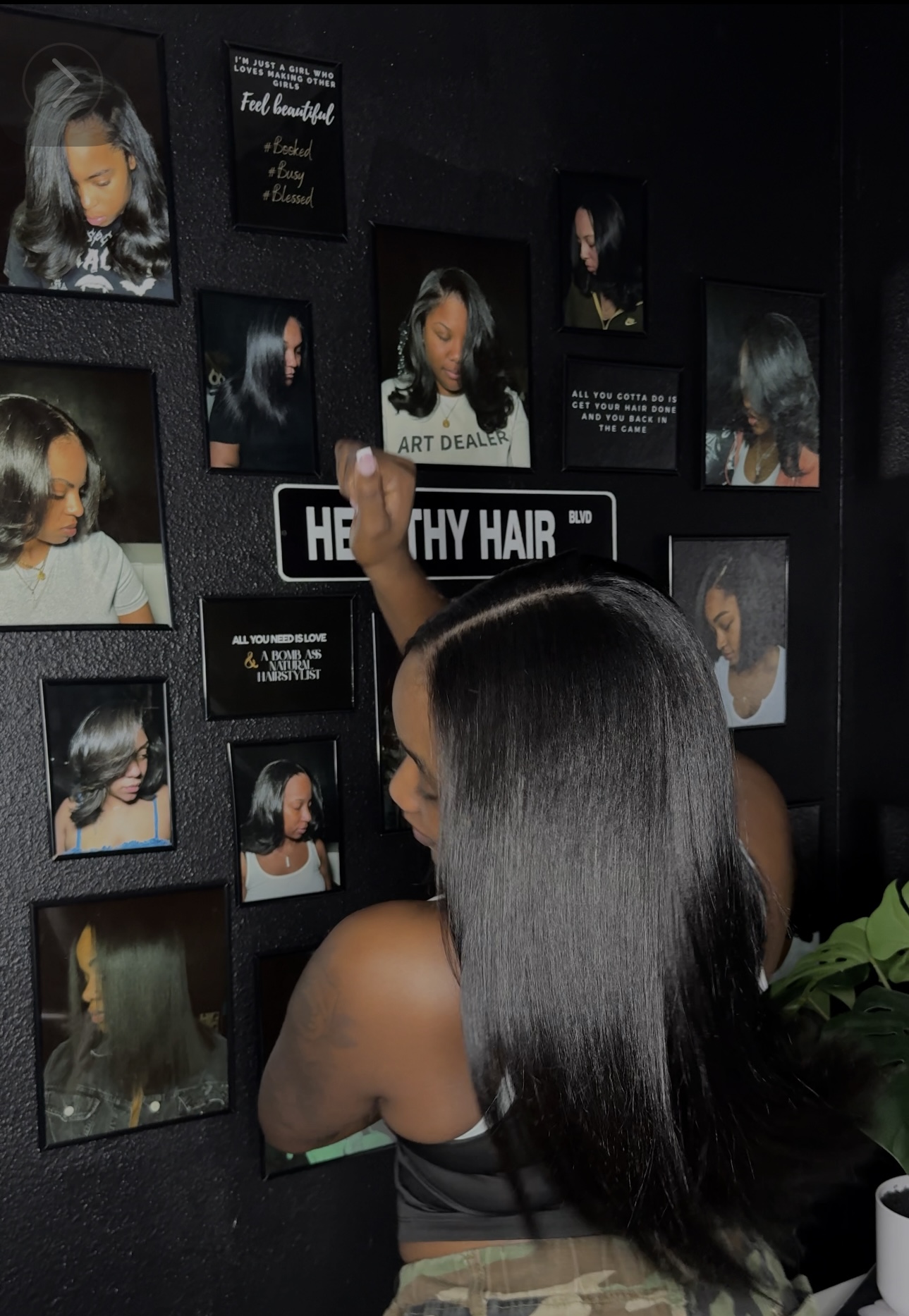 Woman with long black hair reaching for a photo on a wall display of various hair images, some with text. Word 'HEALTHY HAIR' visible.
