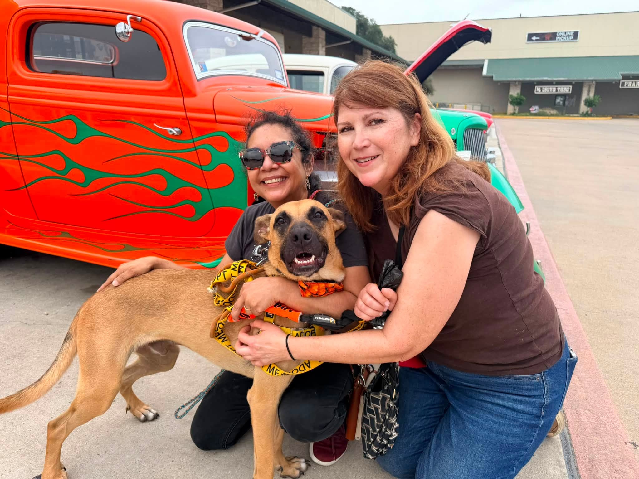 Two women and a dog outdoors near a colorful vintage car, smiling and hugging the dog.