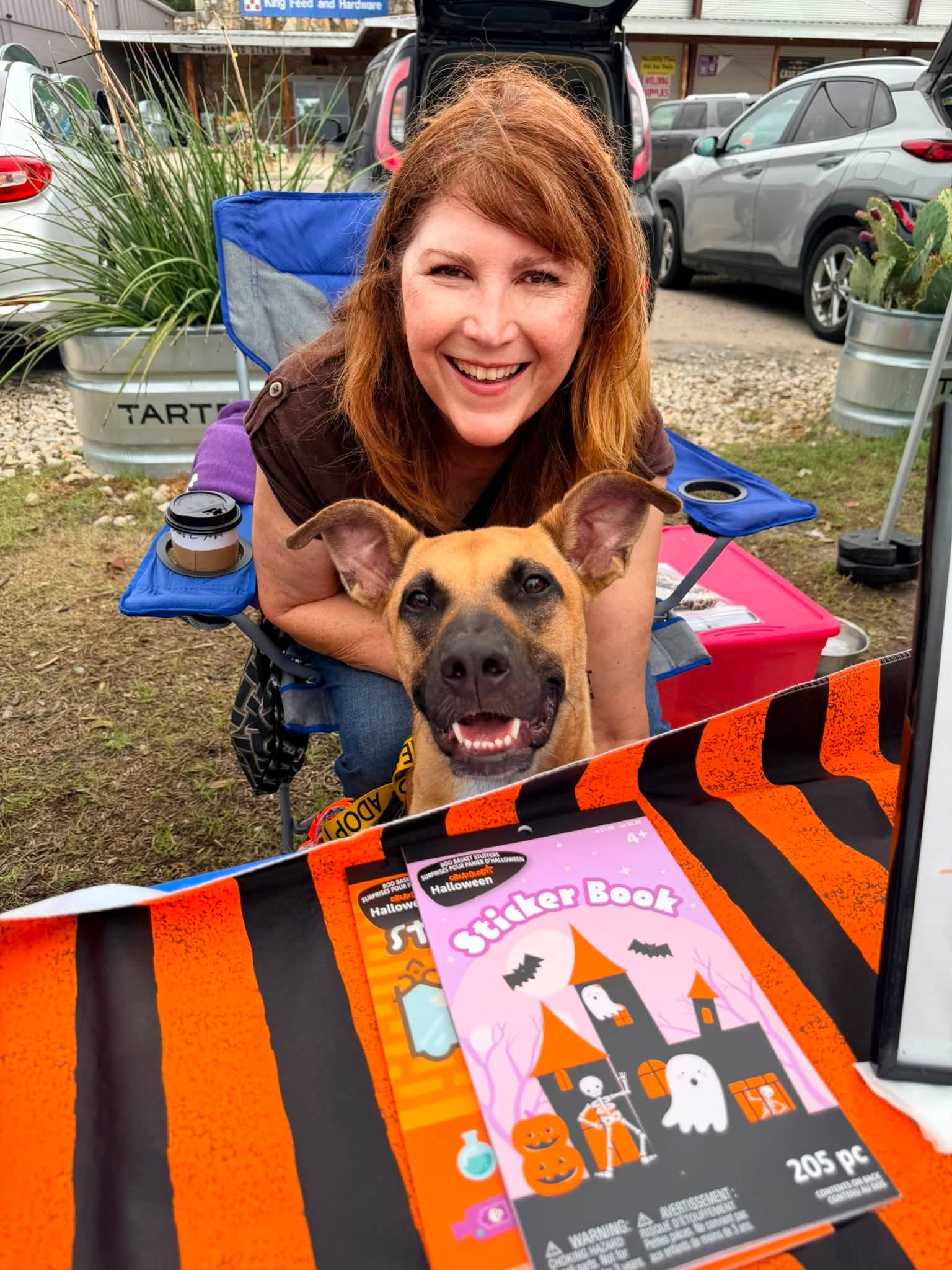 Woman with red hair smiling behind a dog, outdoors with cars and plants in background.
