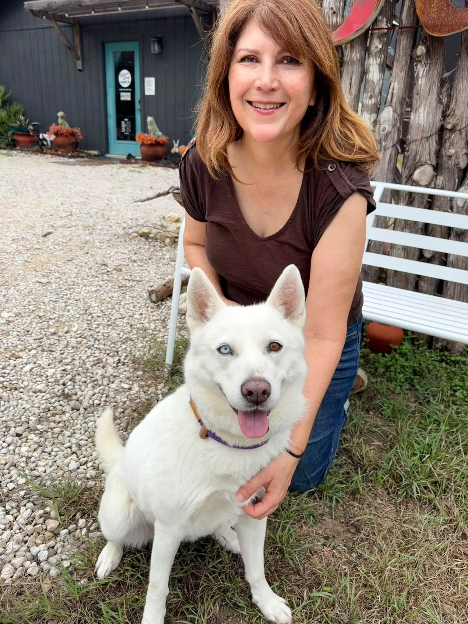 Woman kneeling outdoors with a white dog, smiling, in front of a building and a white bench.