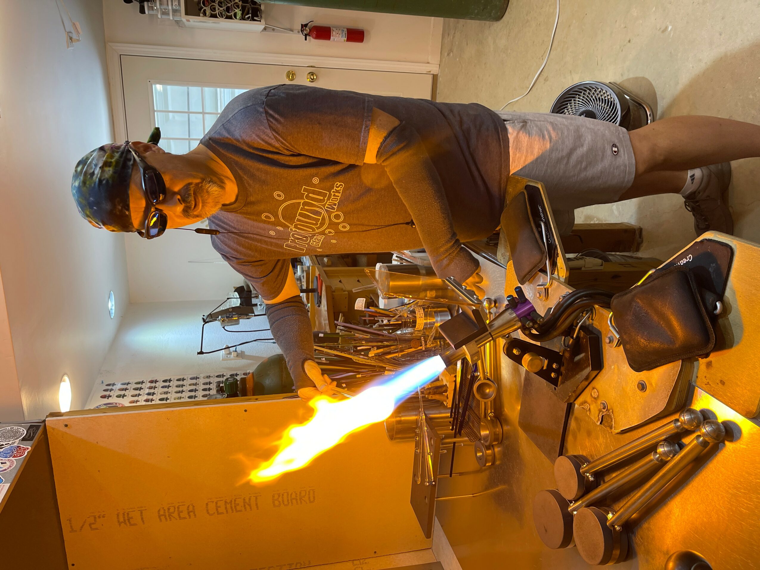 Person wearing safety glasses and a cap, working with a torch producing a flame, in a workshop setting.
