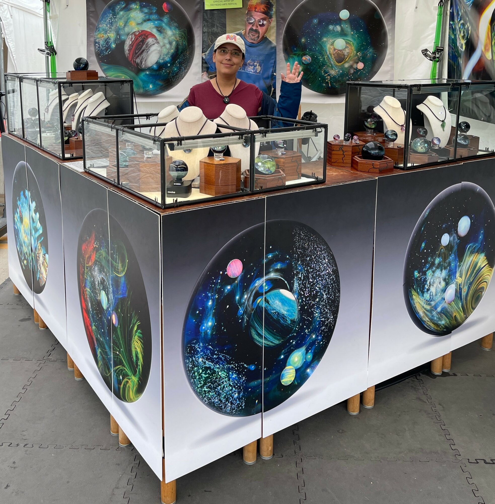 Person standing behind a display booth with space-themed artwork and merchandise, colorful space images on the booth's sides.