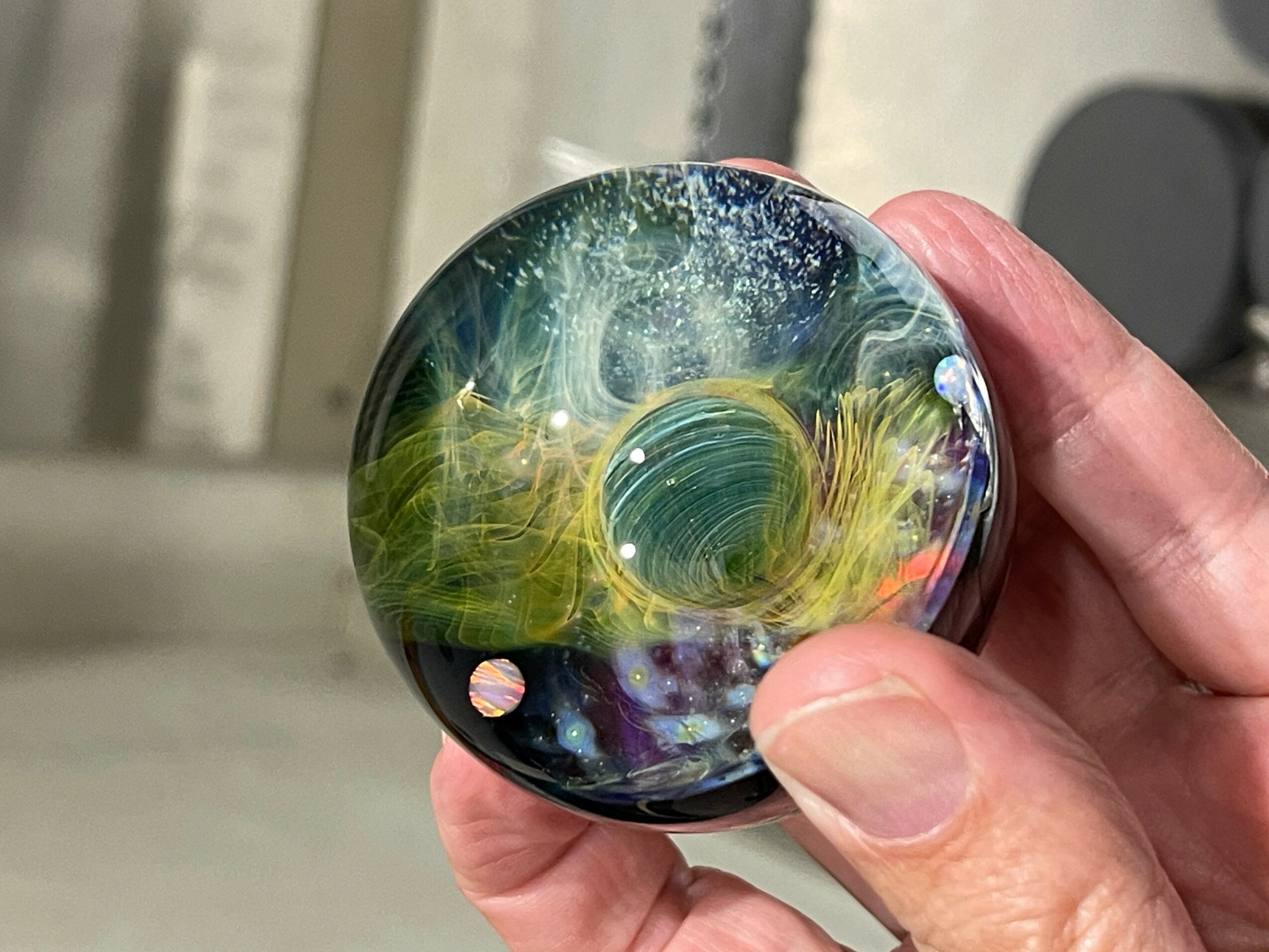 Hand holding a glass marble with colorful swirls and patterns inside, background blurred.