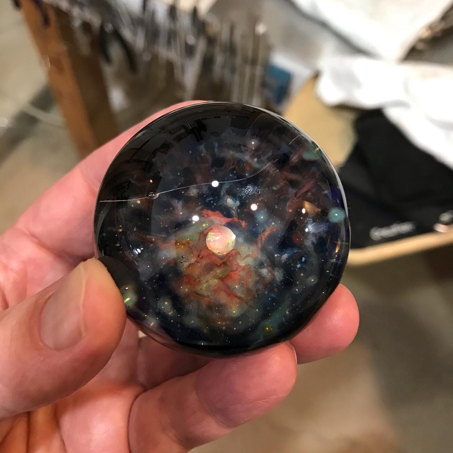 Hand holding a round object with a galaxy design, background shows shelves and fabric items.