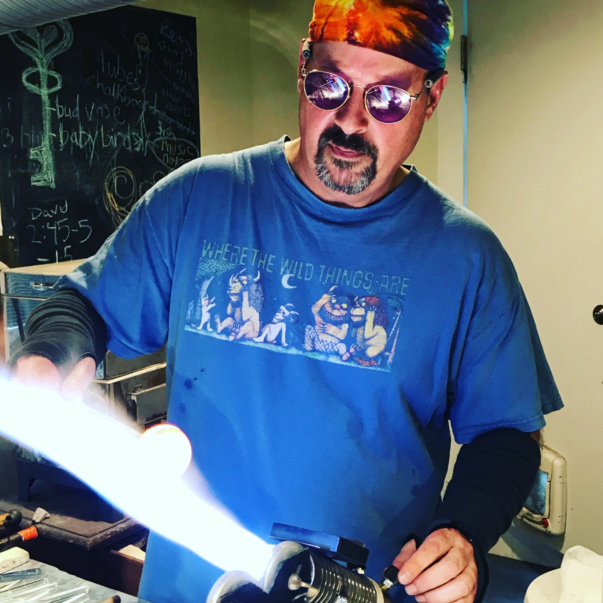 Man wearing sunglasses and a colorful bandana, working with a flame, in a room with a chalkboard in the background.