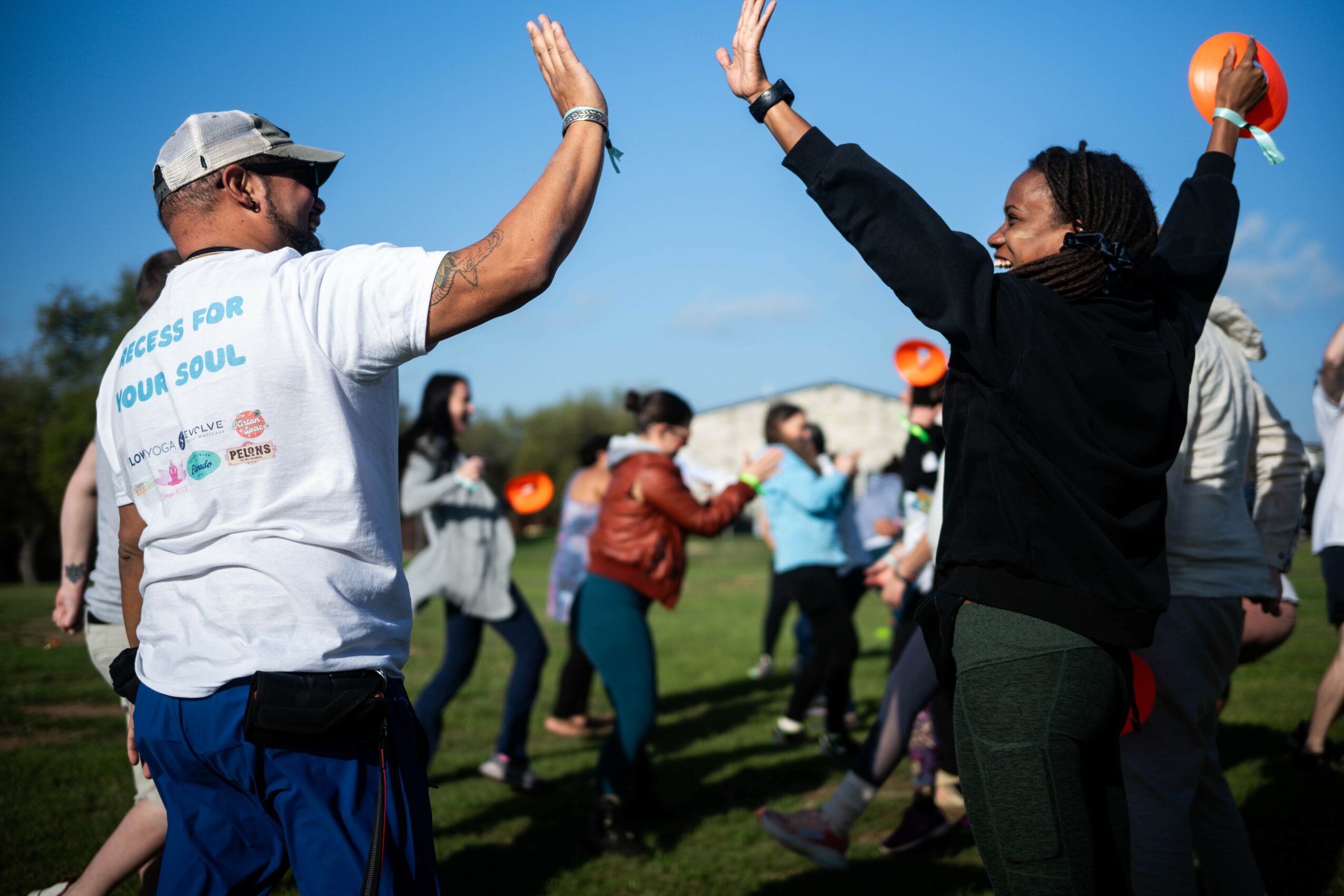 People high-fiving outdoors on a sunny day, some holding orange balls, with a clear blue sky and trees in background.