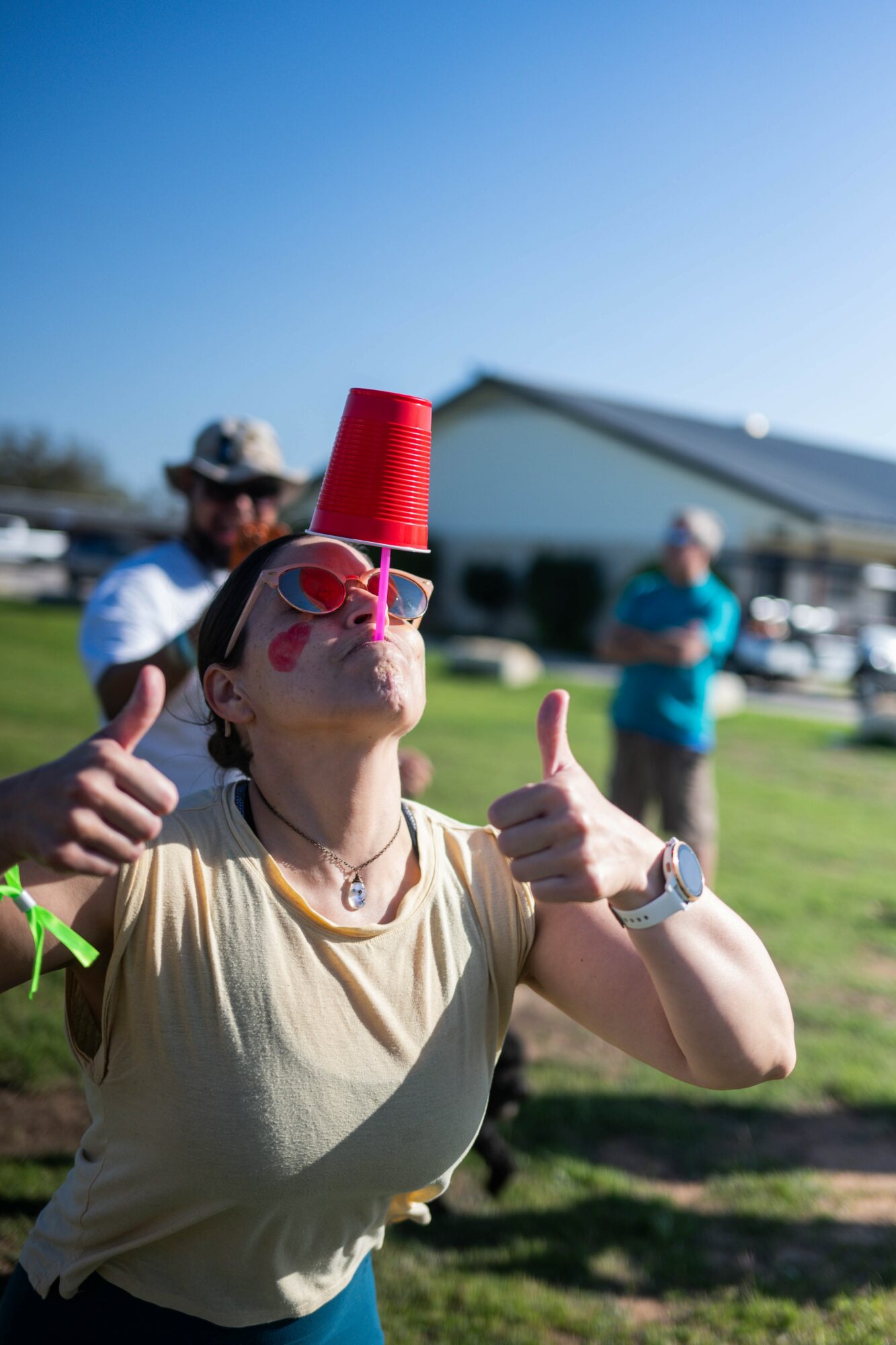 Person balancing a red cup on their head, giving thumbs up outdoors with a building and people in background.