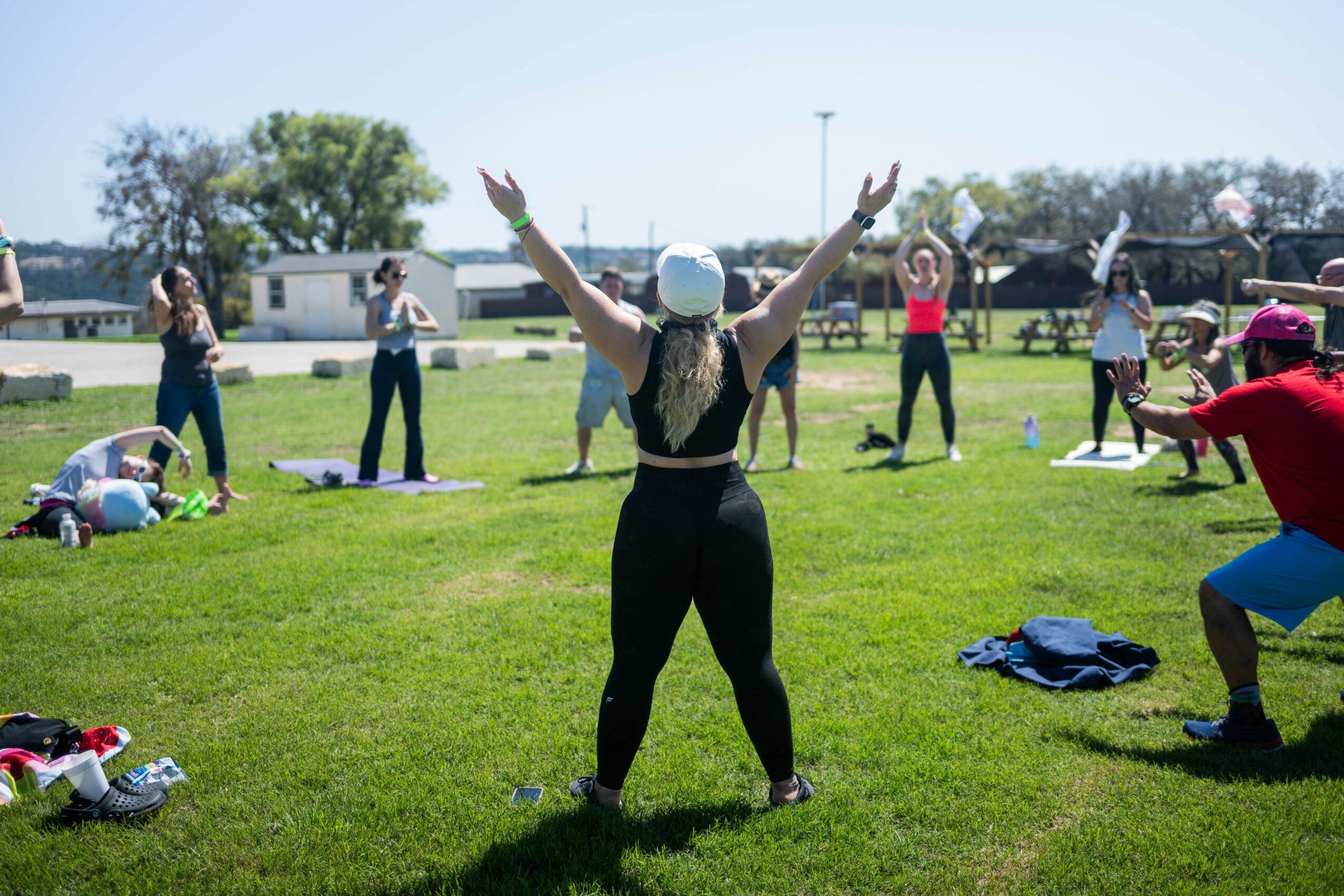 Group of people participating in outdoor exercise class on grass, with instructor facing away, arms raised, and others following.