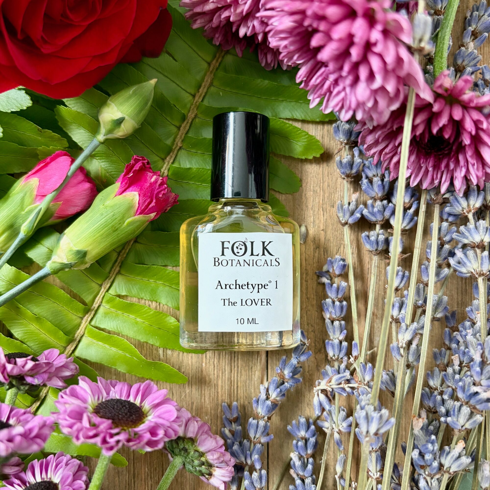 Small perfume bottle labeled 'FOLK BOTANICALS Archetype The LOVER' surrounded by colorful flowers on a wooden surface.