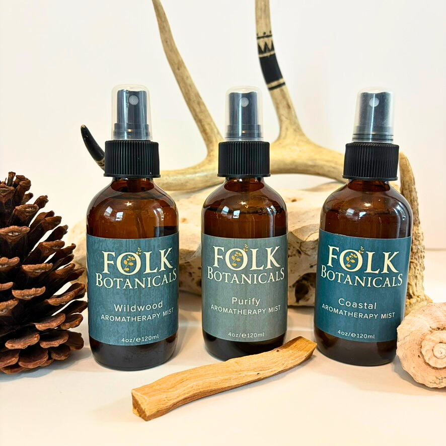Three bottles of folk botanicals aromatherapy mist with pinecones, antler, and wood pieces in background, on a white surface.