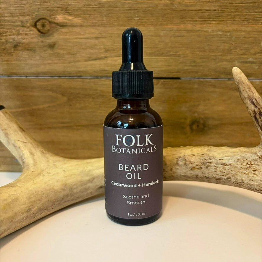 Small dark brown bottle with a black dropper cap labeled Folk Botanicals Beard Oil, placed on a light surface with wooden background.