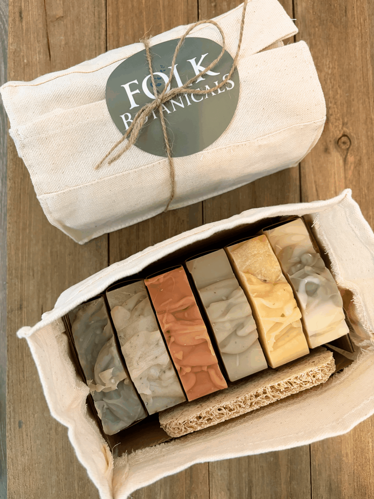 Open box of assorted soap bars with a cloth bag labeled 'FOLK & BOTANICS' on wooden surface.
