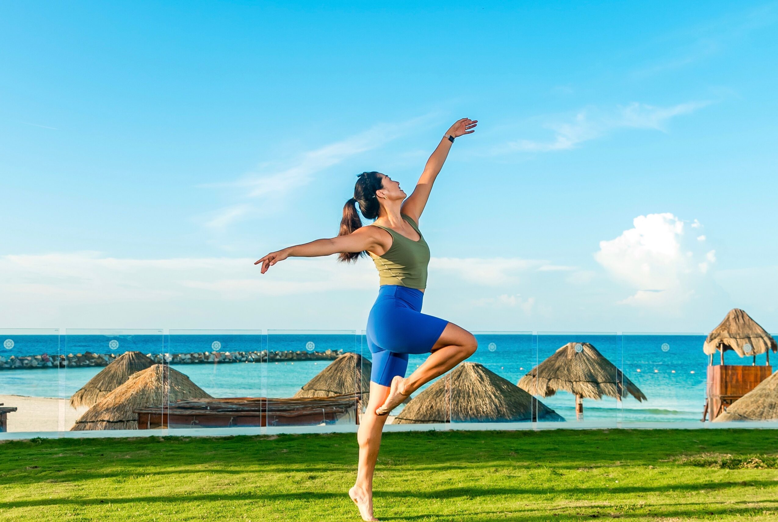 Person balancing on one leg with arms raised on grass beside a beach and turquoise water.