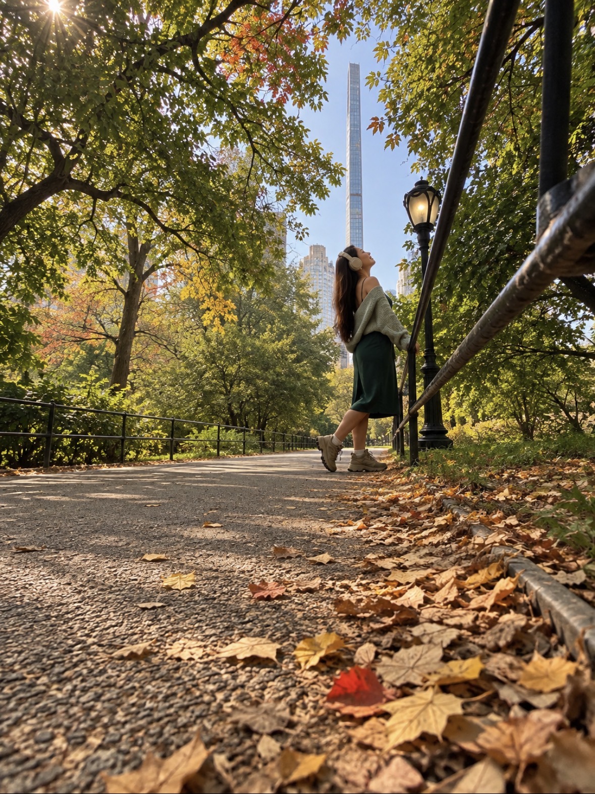 A person walks along a leaf-covered park path beneath autumn trees, with a lamppost and city buildings in the distance.