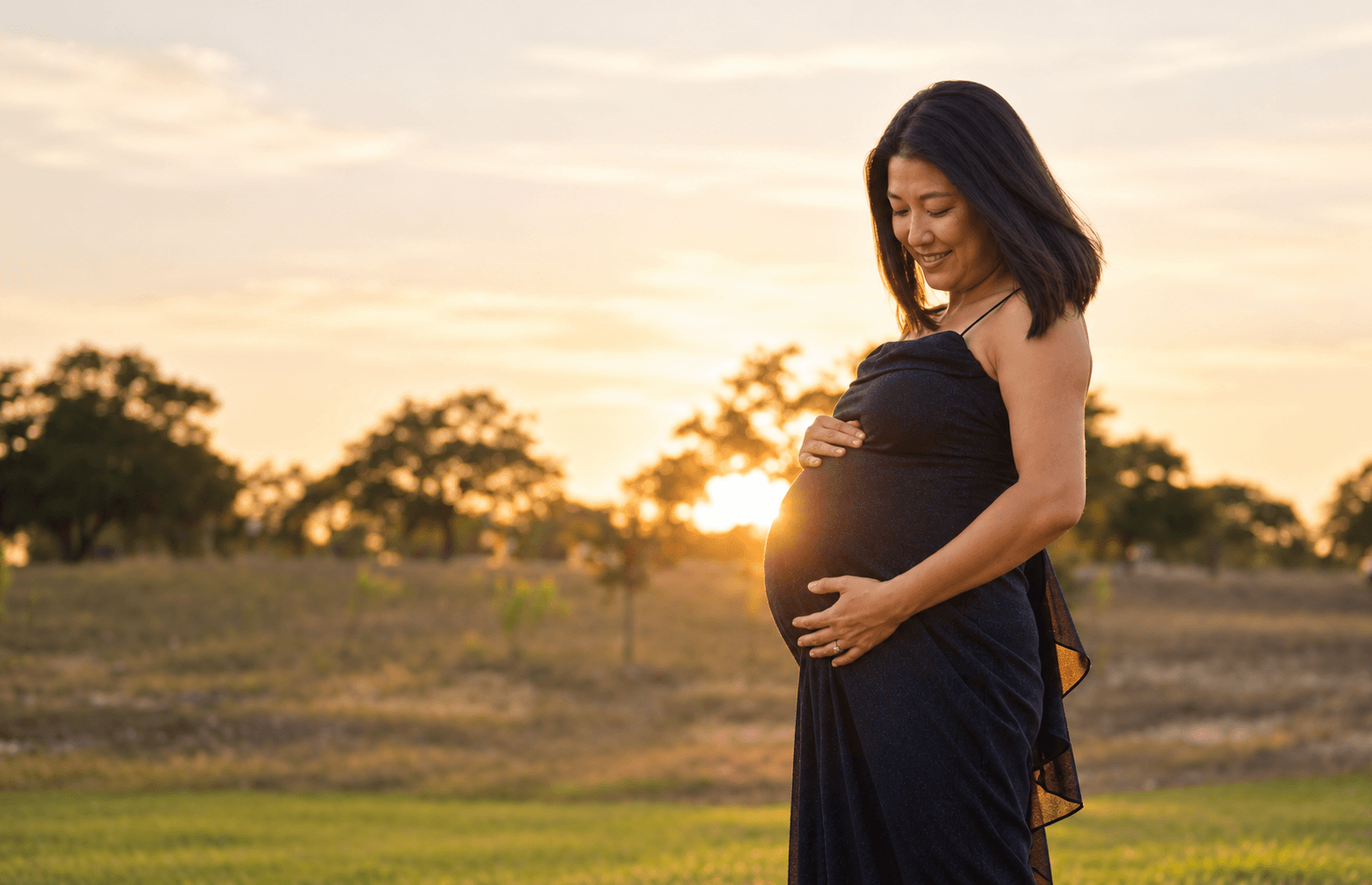 A pregnant person in a dark dress stands outdoors at sunset, holding her belly.