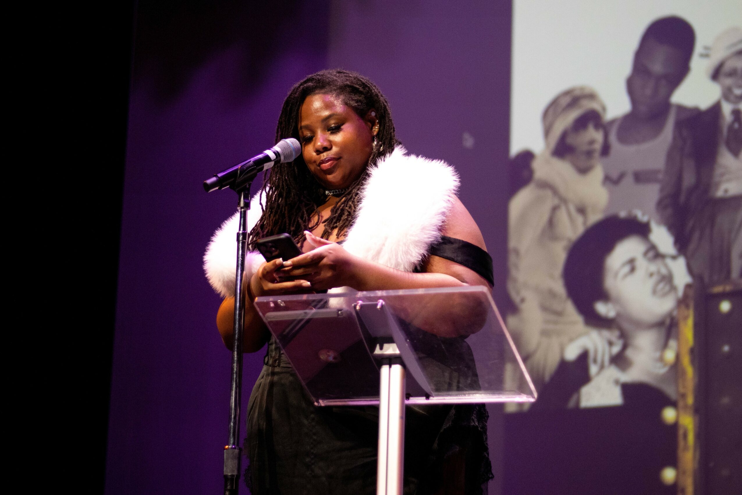 Woman with long hair and white fur stole speaking into a microphone on stage, holding a phone, with a black and white photo in background.