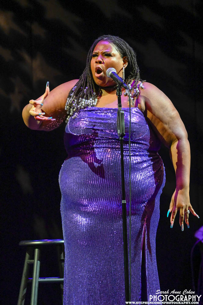 A woman in a purple sequined dress sings into a microphone on stage, with a dark background and a stool nearby.