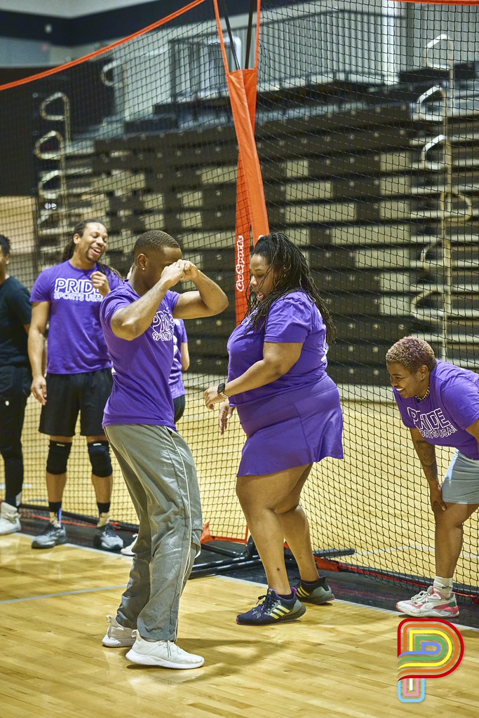 Group of people in purple shirts standing on a gymnasium floor near a volleyball net, some smiling and talking.