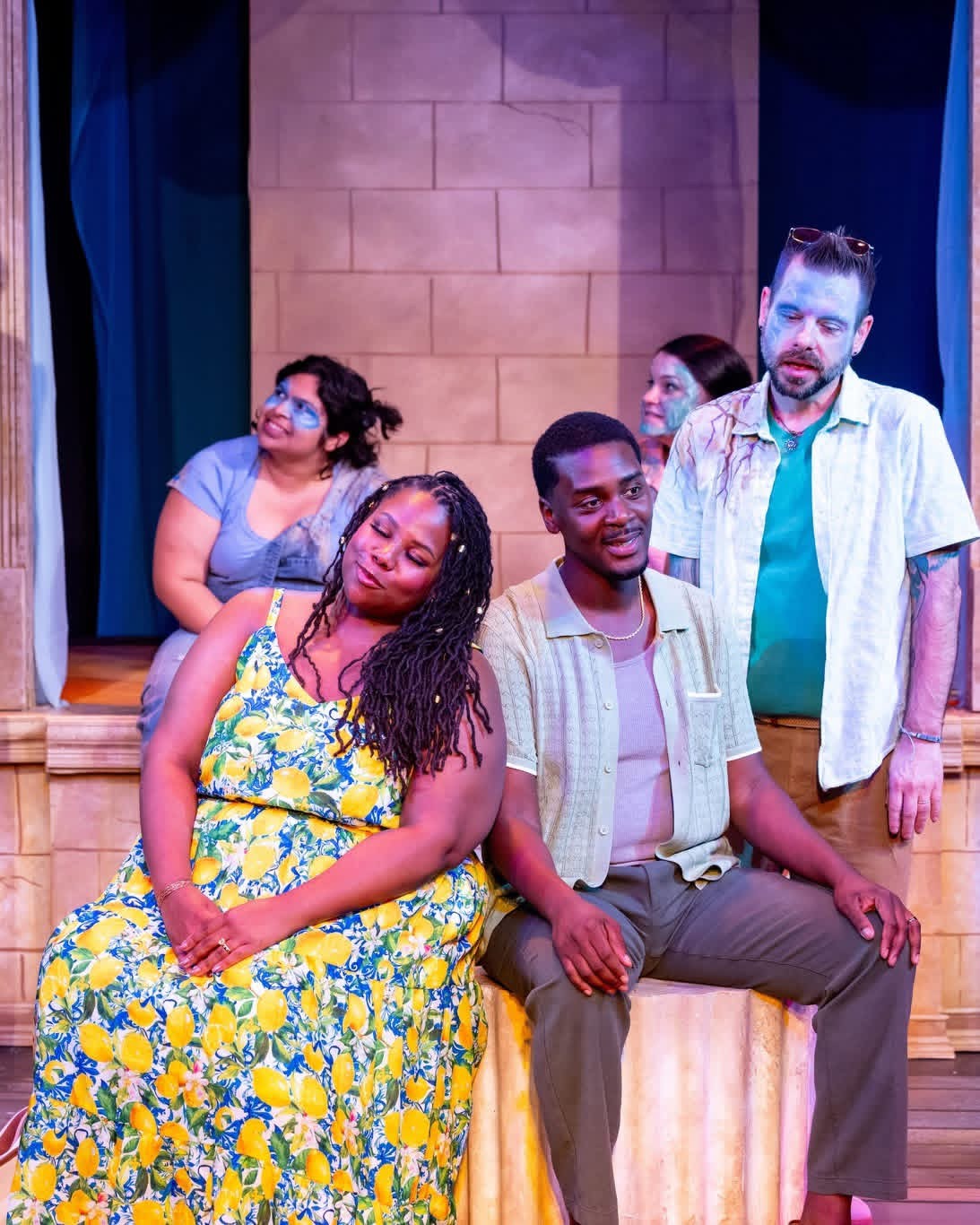 Four people on stage, two sitting and two standing, with a brick wall background and curtains on sides.