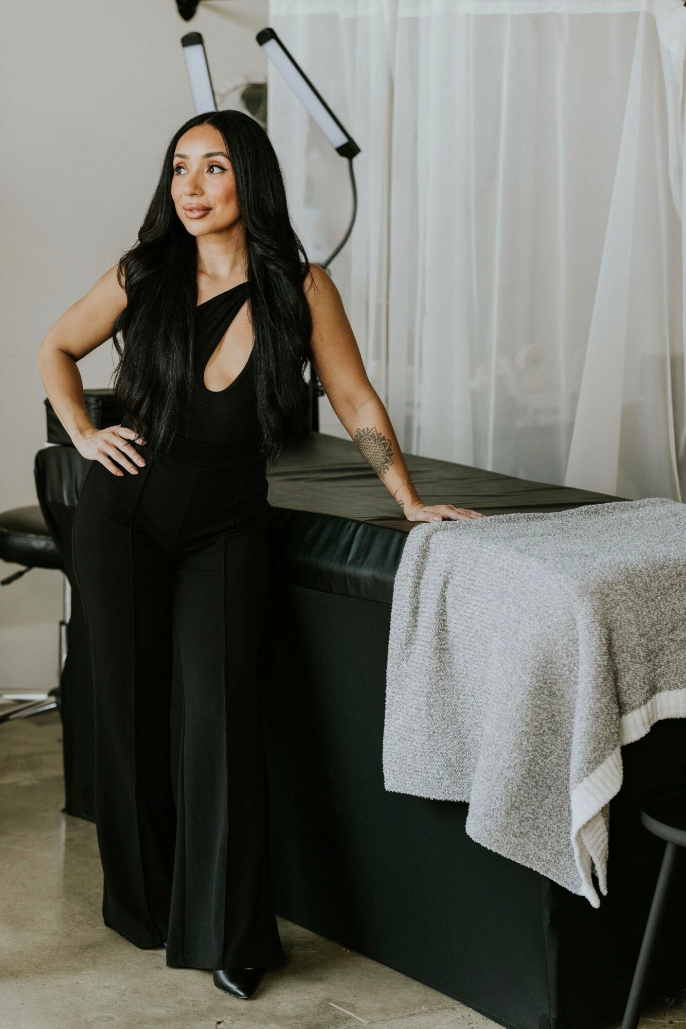 Woman in black outfit standing next to a massage table with a gray blanket, in a room with white curtains.