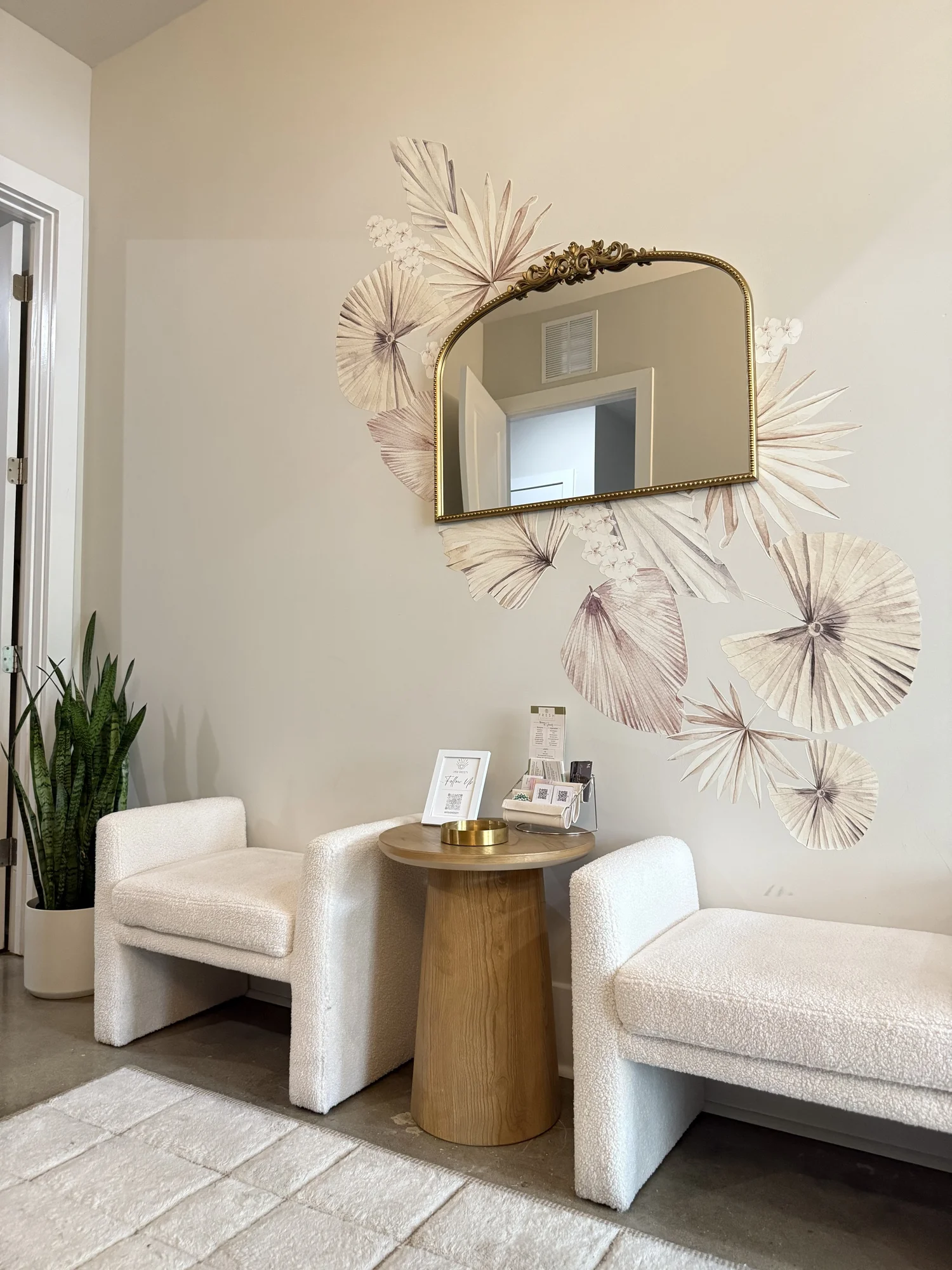 Interior corner with two white armchairs, a round wooden table, a mirror, and decorative wall art resembling dandelions.