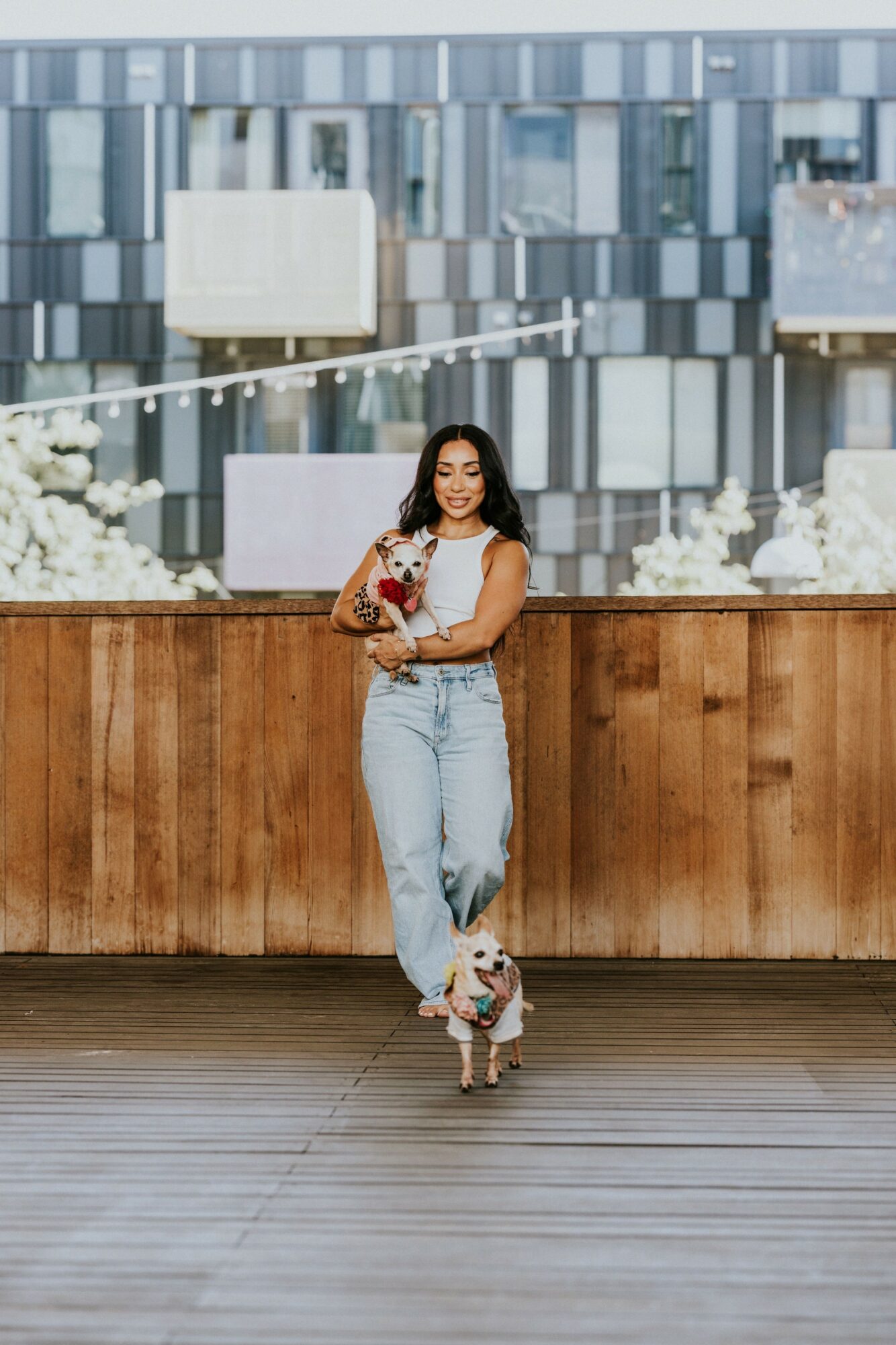 Young woman standing with a dog in front of a wooden wall, city buildings in background, holding a drink, smiling.