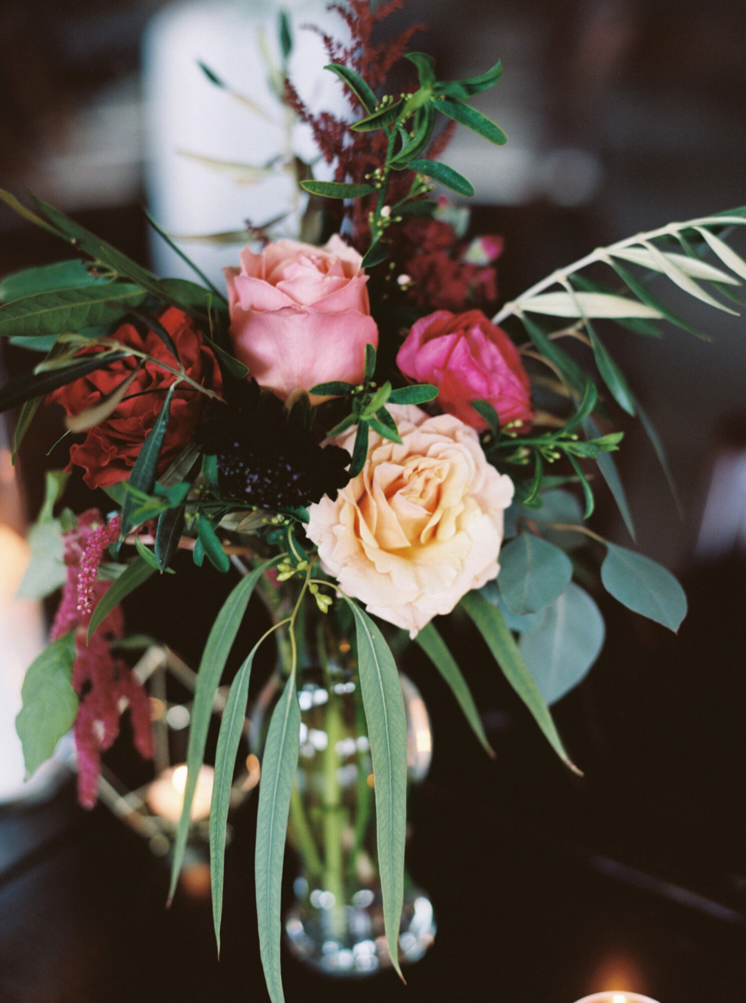 A floral arrangement with pink, peach, and dark red flowers, surrounded by green leaves and foliage in a glass vase.
