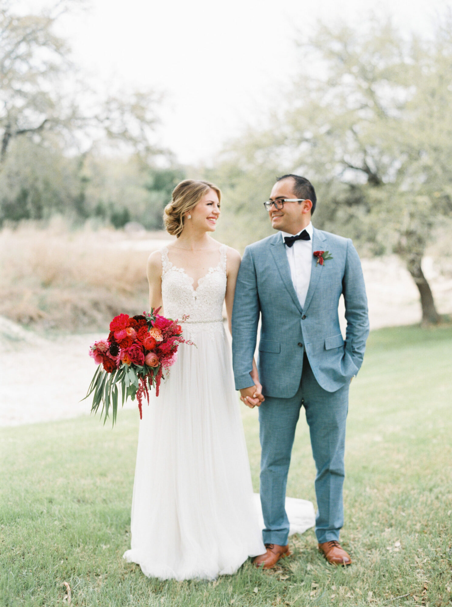 A bride and groom holding hands outdoors, the bride holding a bouquet of red flowers, trees in the background.