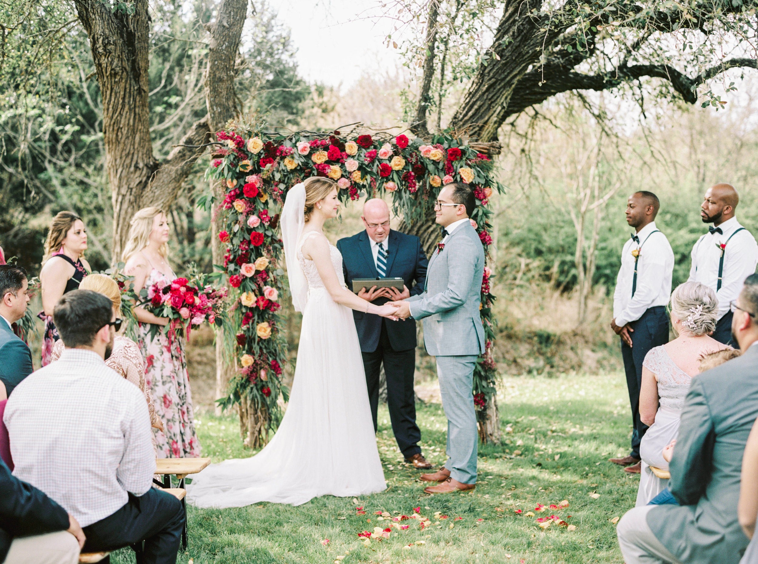 Bride and groom holding hands during outdoor wedding ceremony with floral arch, surrounded by guests and trees.