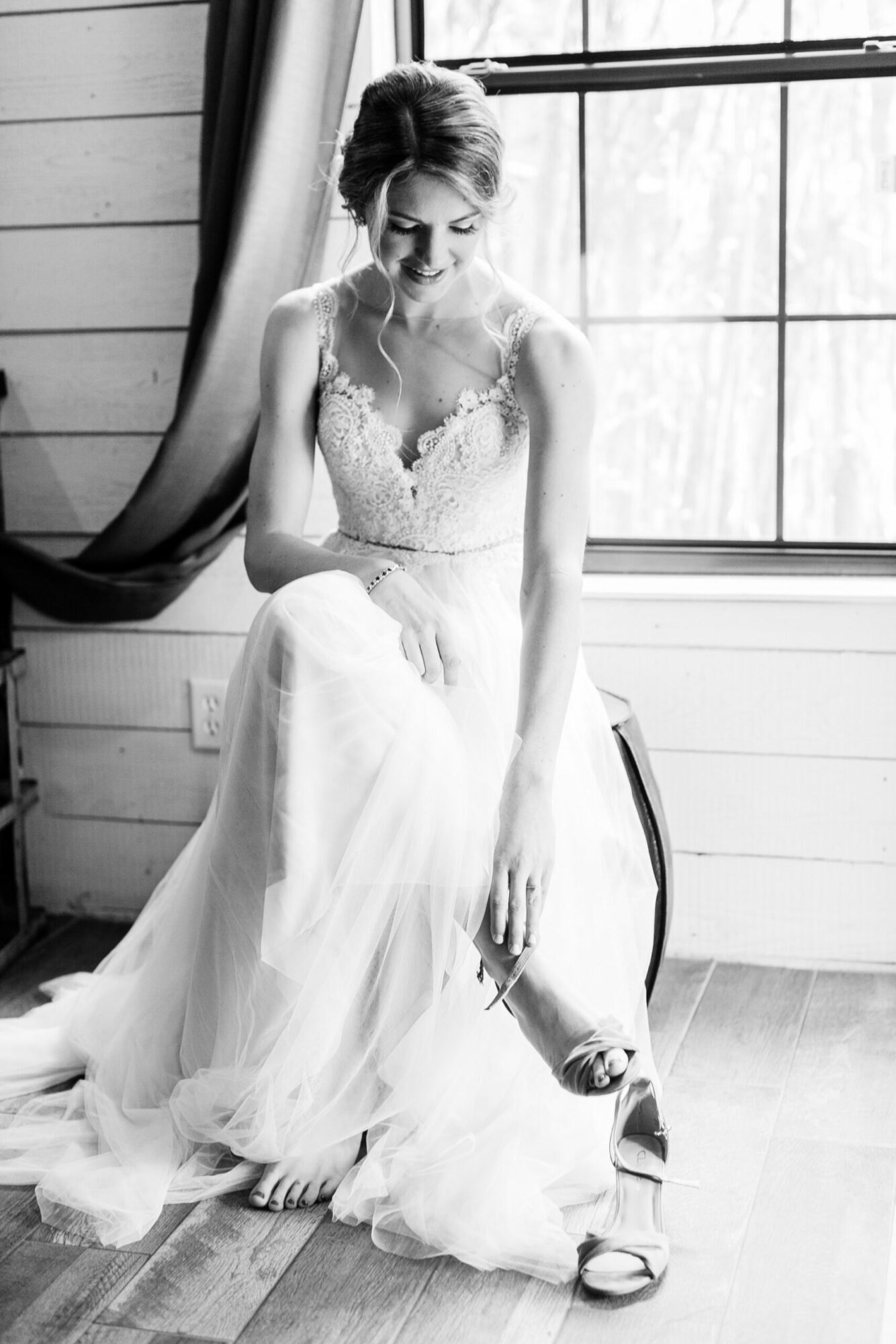 Woman in wedding dress sitting by window, holding a shoe, in a room with wooden walls and large window.