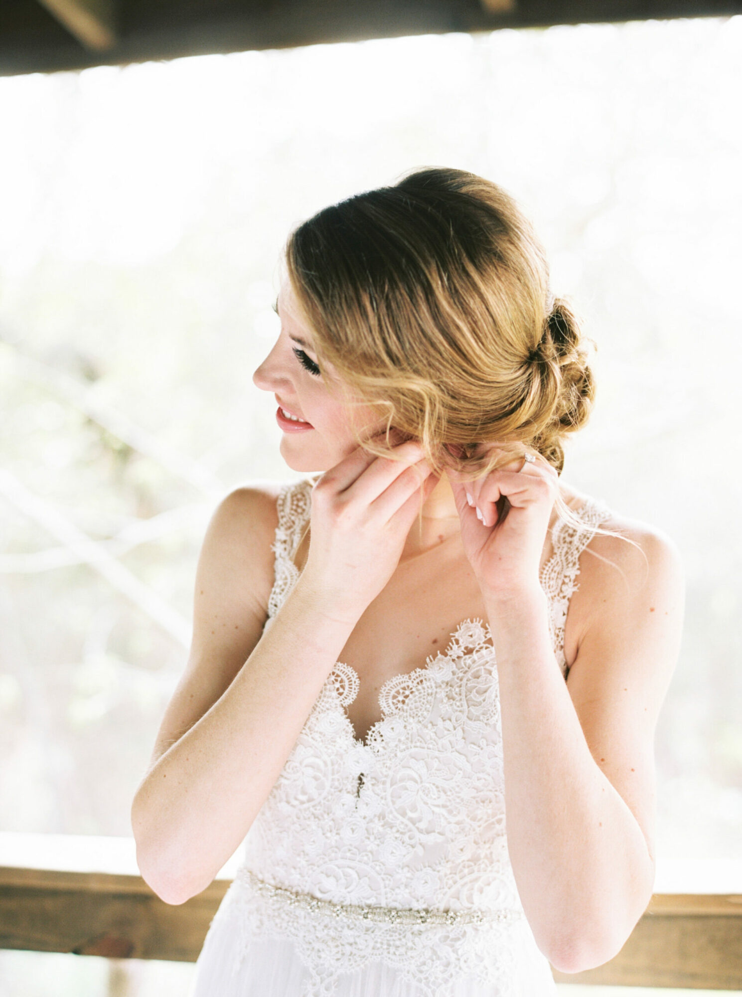 Woman with blonde hair in a lace dress adjusting her earring, smiling, near a window with natural light.