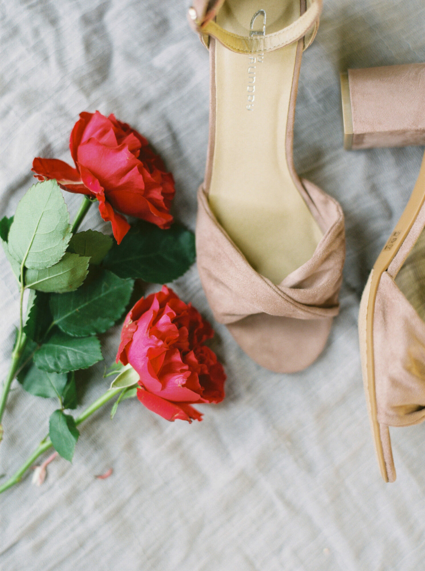 Two red roses with green leaves, a beige high-heeled shoe, and a pink box on a light fabric surface.