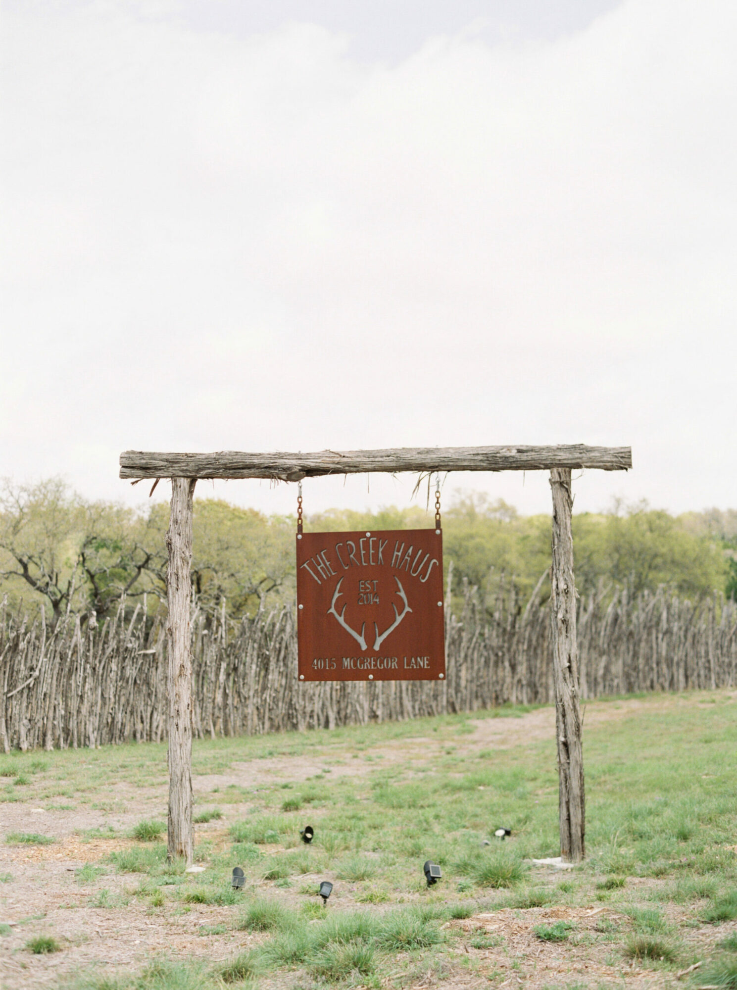 Wooden archway with a hanging sign featuring a deer head silhouette, set in a grassy field with trees in the background.