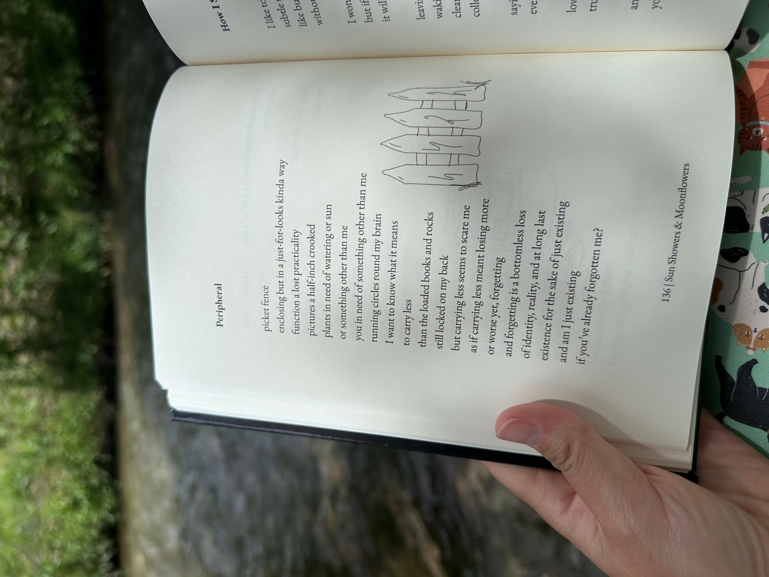 Open book showing text and a line drawing of buildings, held by a person's hand outdoors with greenery and water in background.