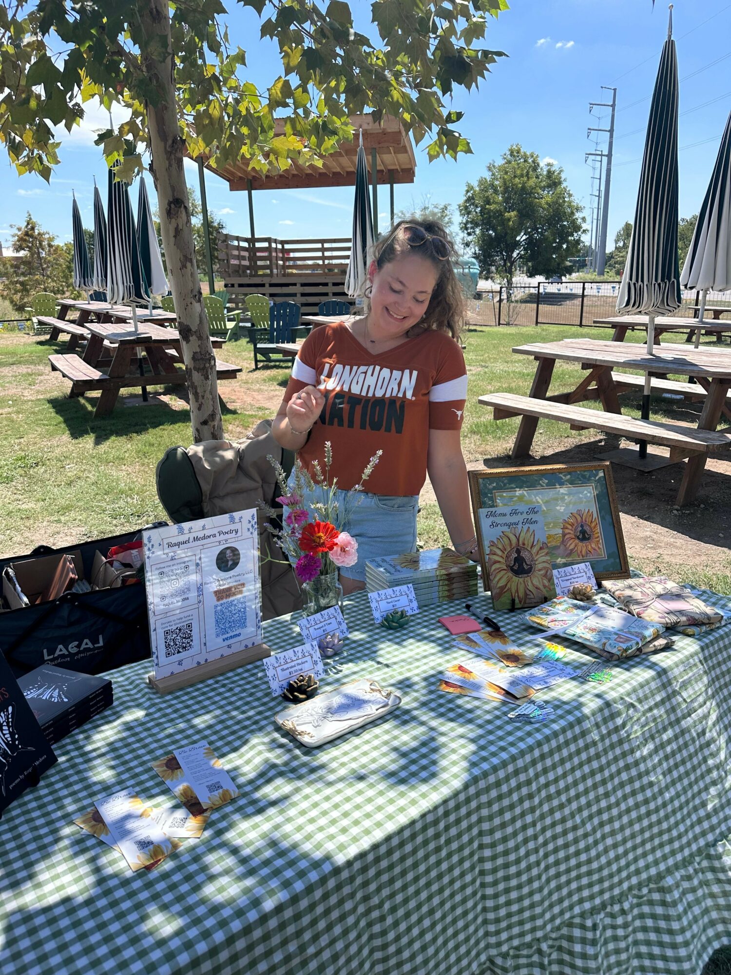 Woman standing at outdoor table with books, flowers, and papers, under a tree with picnic tables and a fence in background.