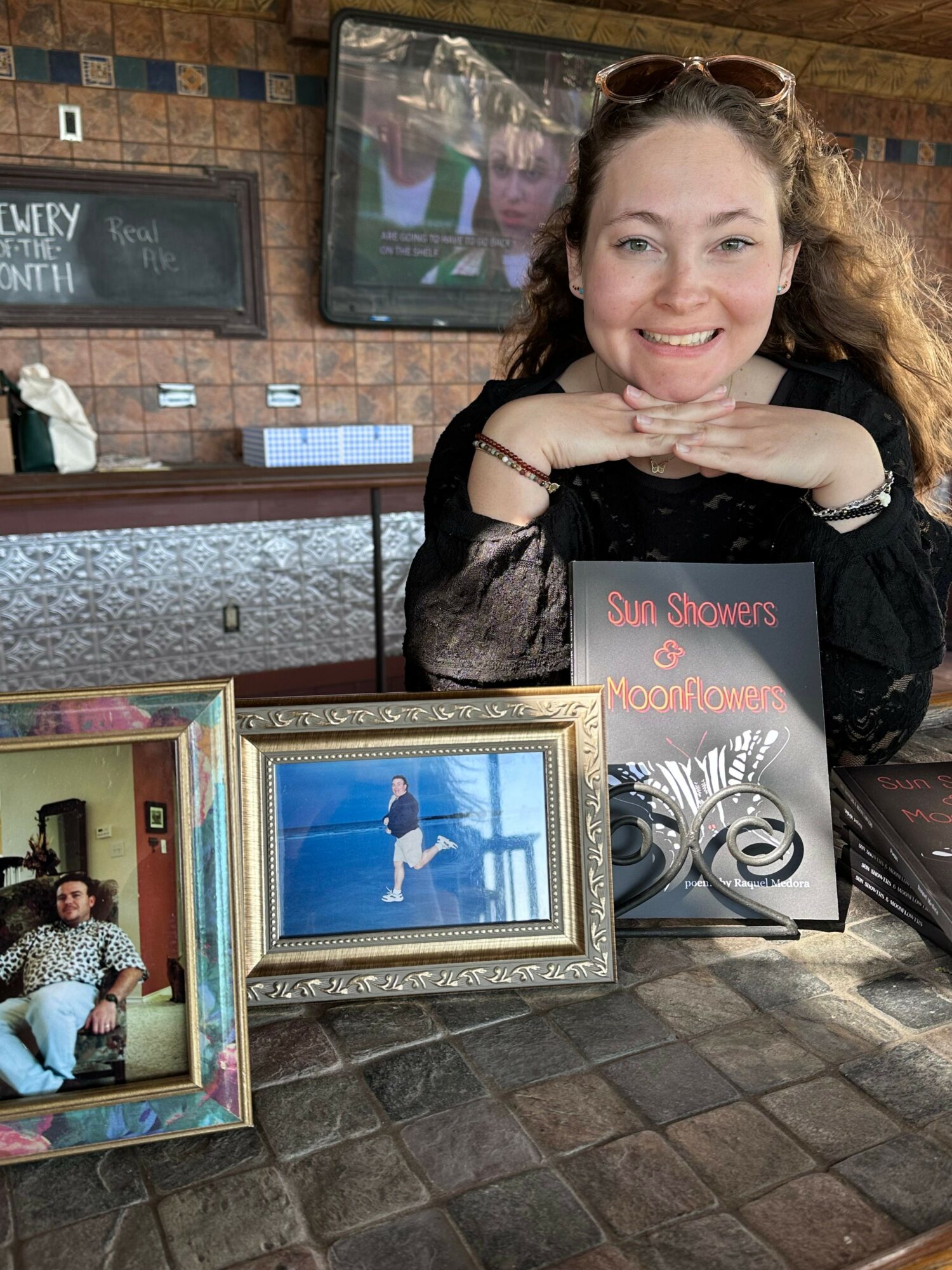 Woman with curly hair lying on a table, smiling, surrounded by framed photos and posters, brick wall background.