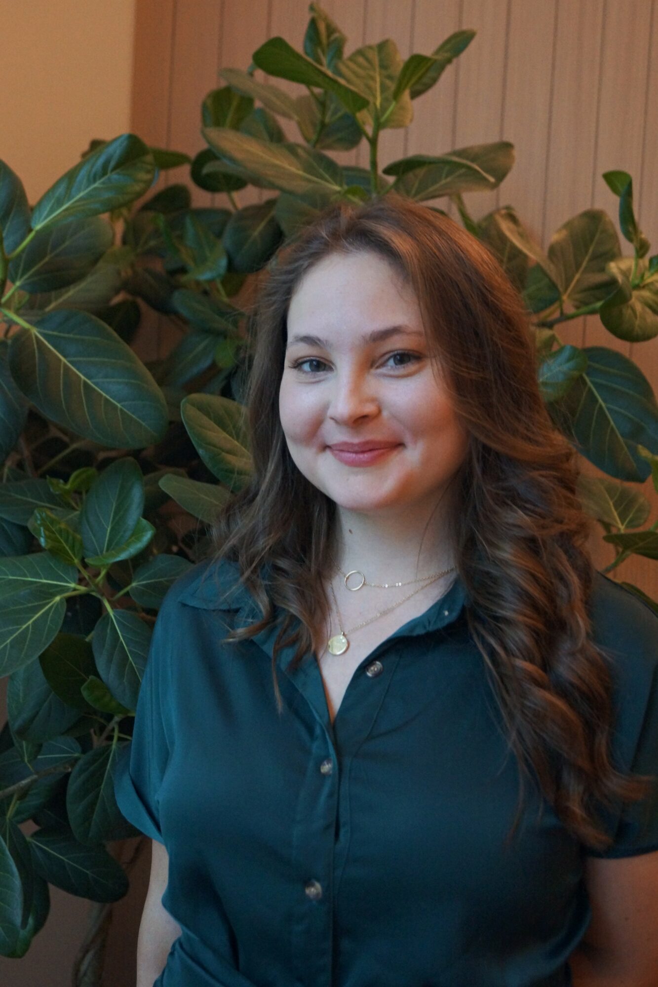 Young woman with long wavy hair smiling, standing in front of large green leaves, wearing a dark shirt.