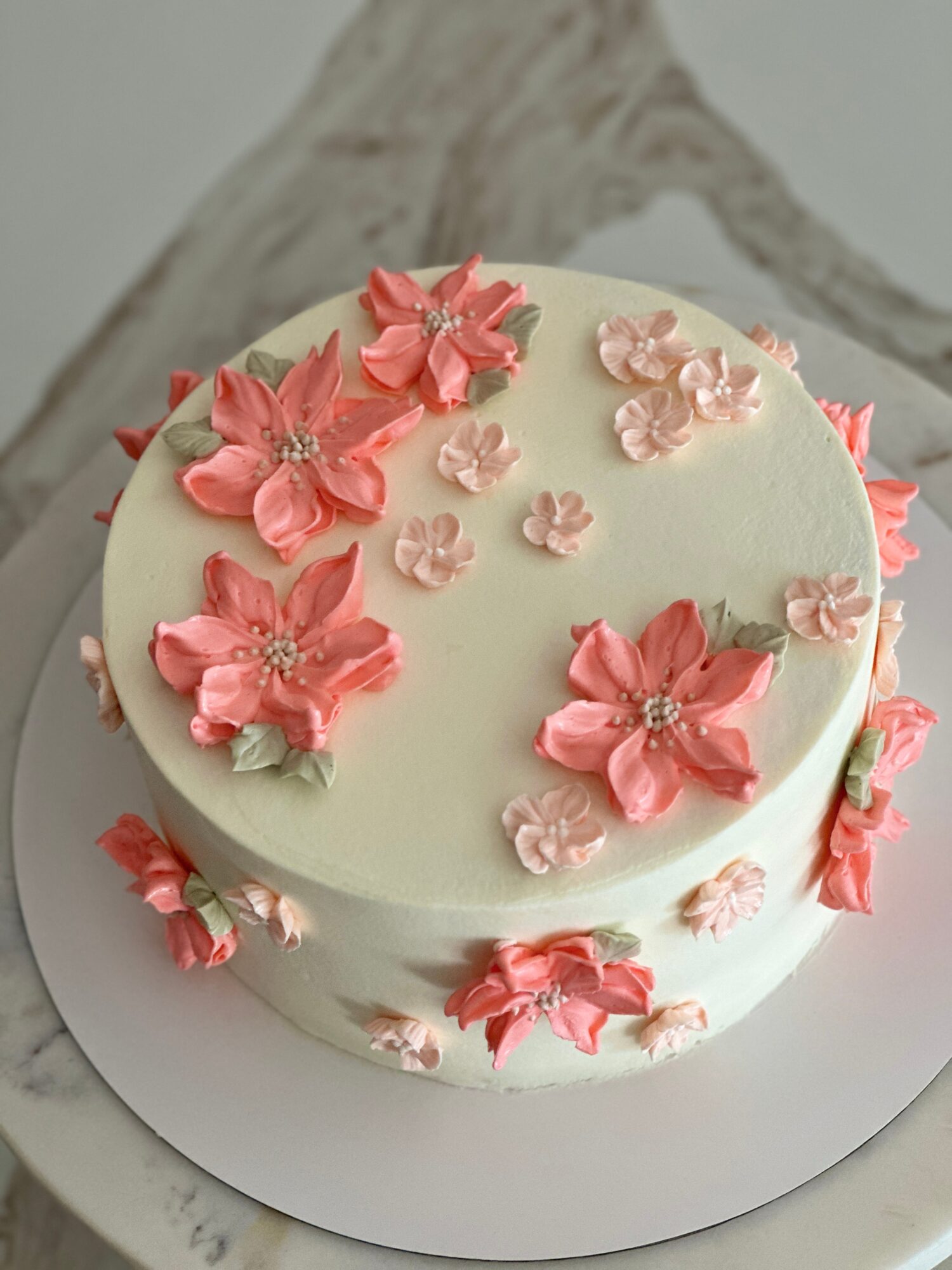 Round white cake decorated with pink flower-shaped frosting and small blossoms on a cake board.