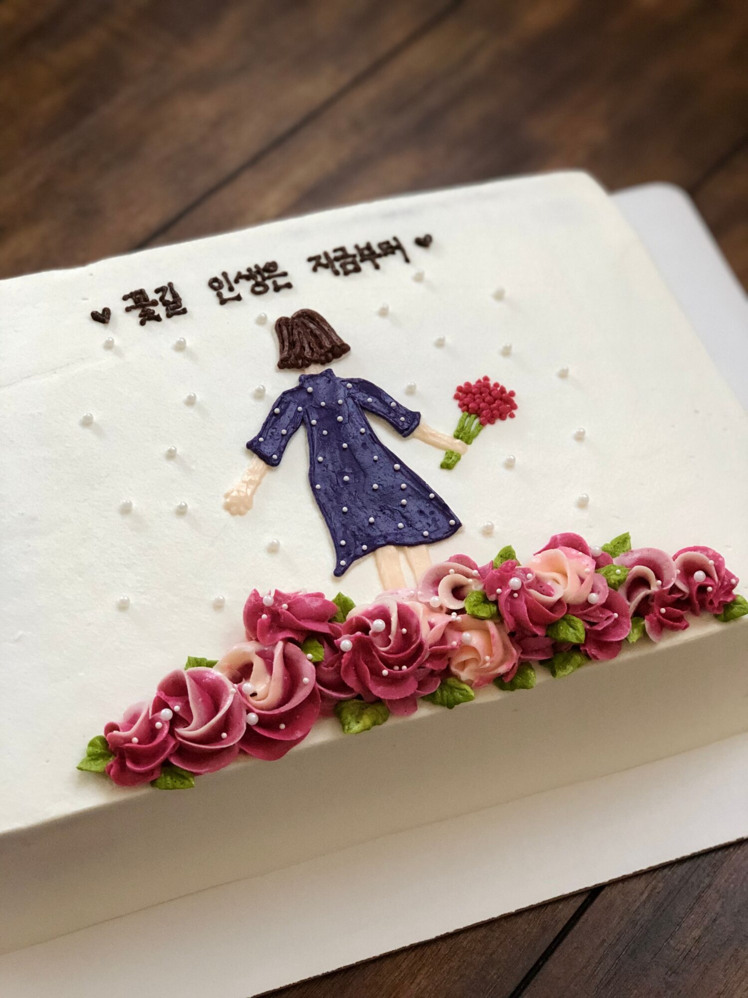 Rectangular cake decorated with Korean writing, a figure holding a flower, and pink floral frosting.
