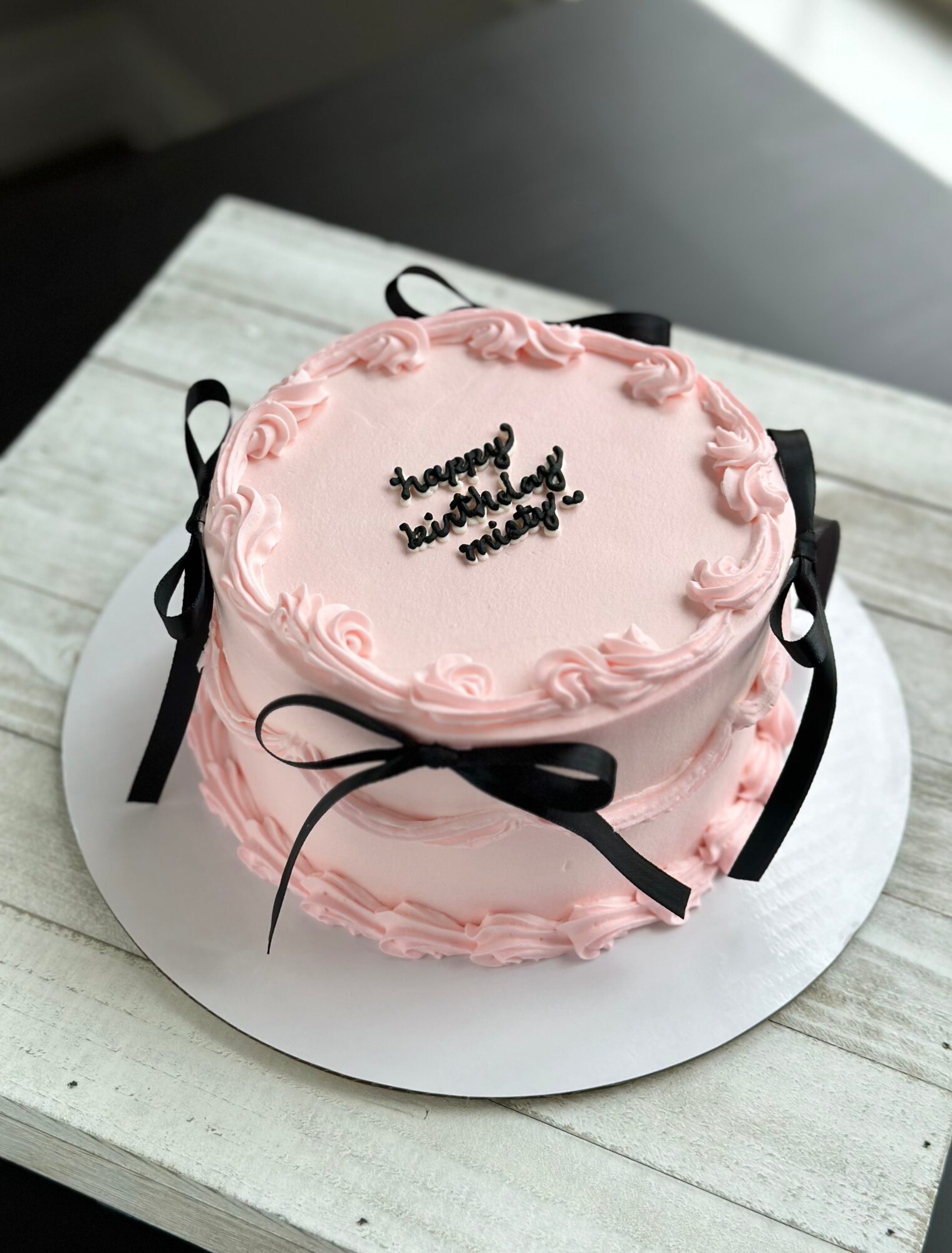 Round pink cake with piped frosting, black ribbon decorations, and writing on top, displayed on a white board.