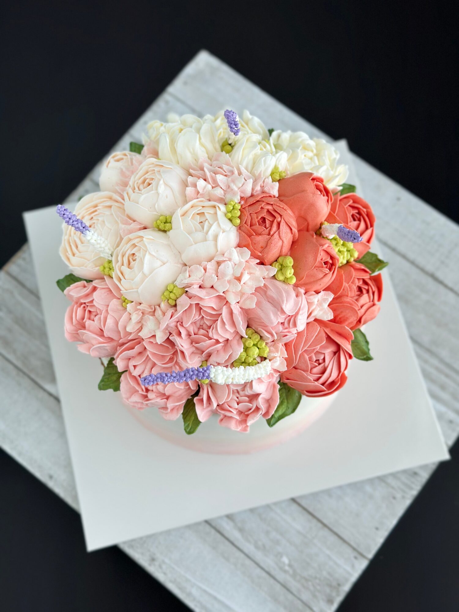 A round bouquet of pink, coral, and white flowers sits on a white square board over a gray wooden surface.