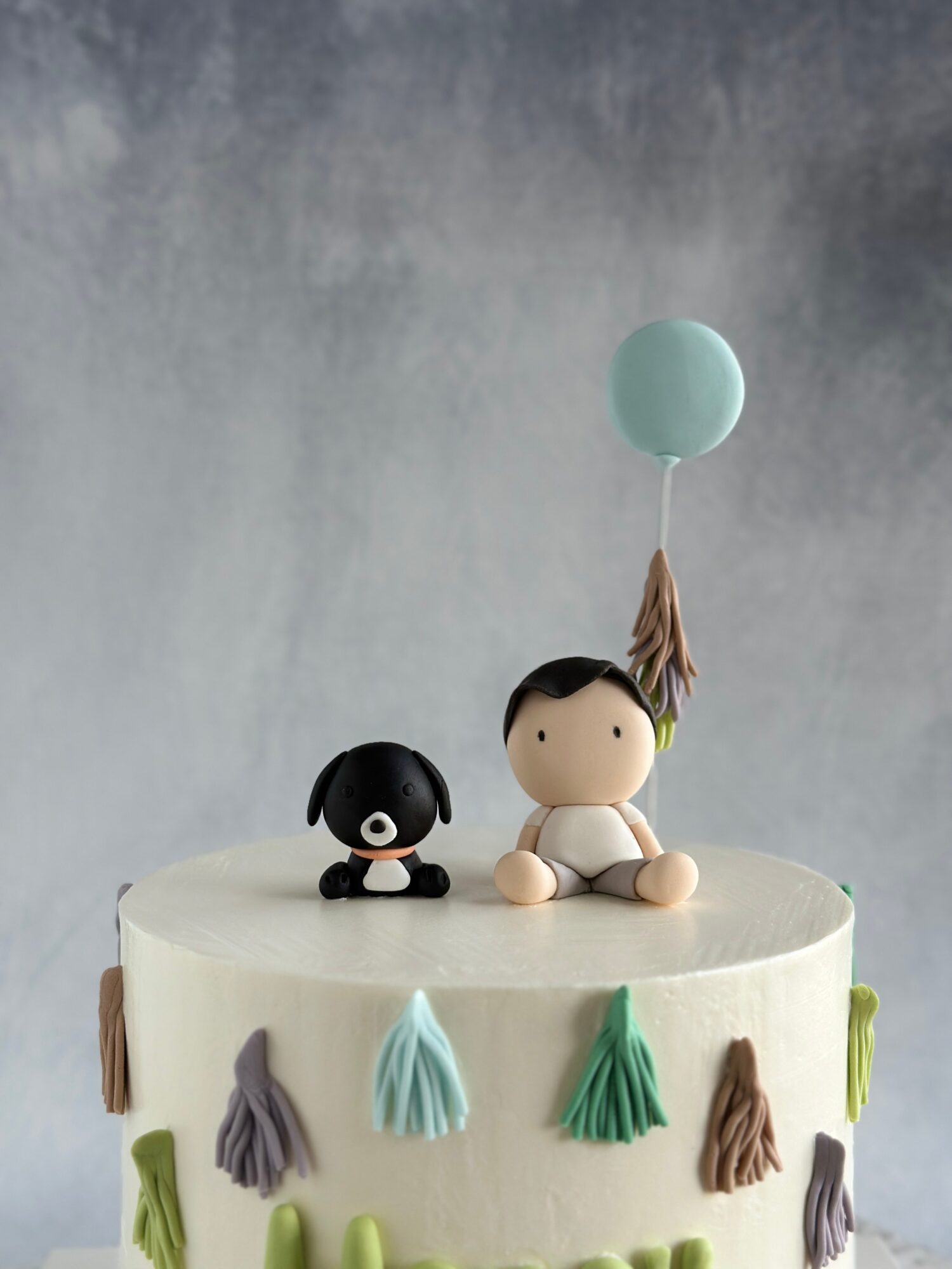 Two small figurines sit on a decorated white cake, with a teal balloon floating above them.