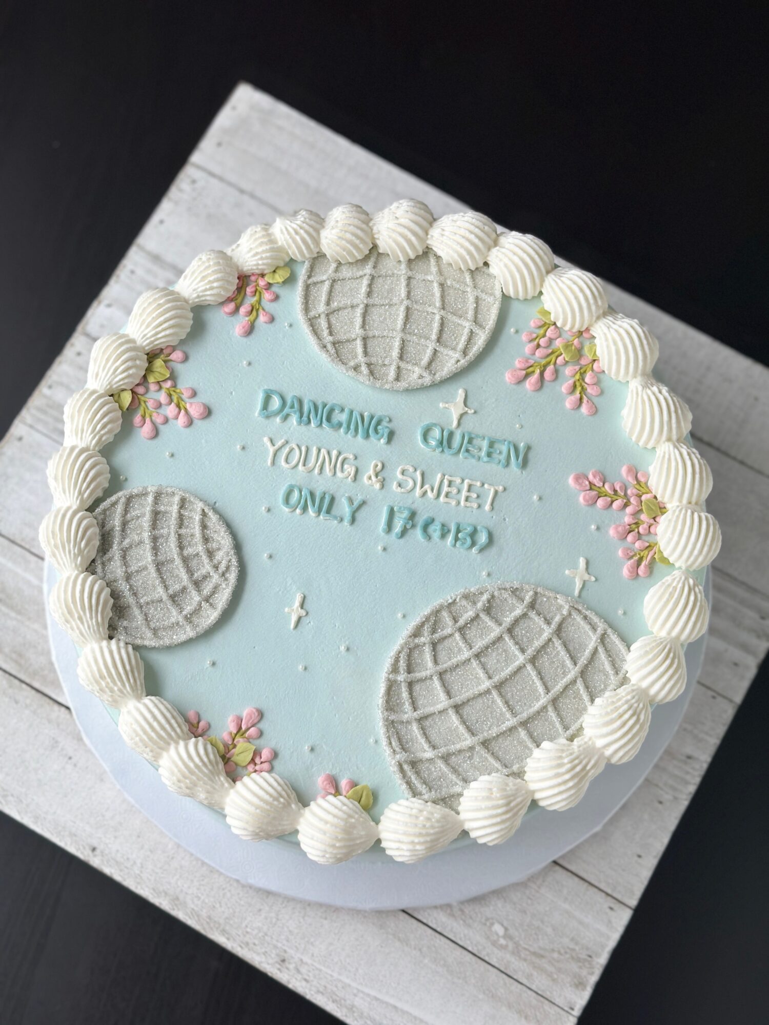 Round blue cake with piped cream borders, shell decorations, flowers, and a written message on a white board.