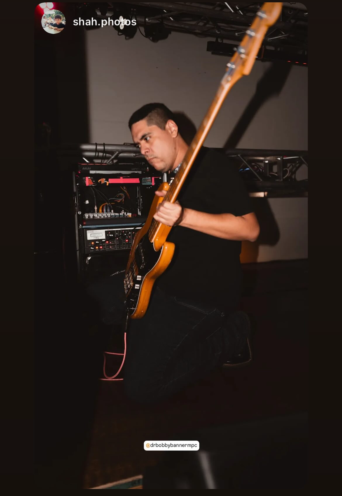 A person plays an electric bass guitar on a dimly lit stage beside audio equipment.