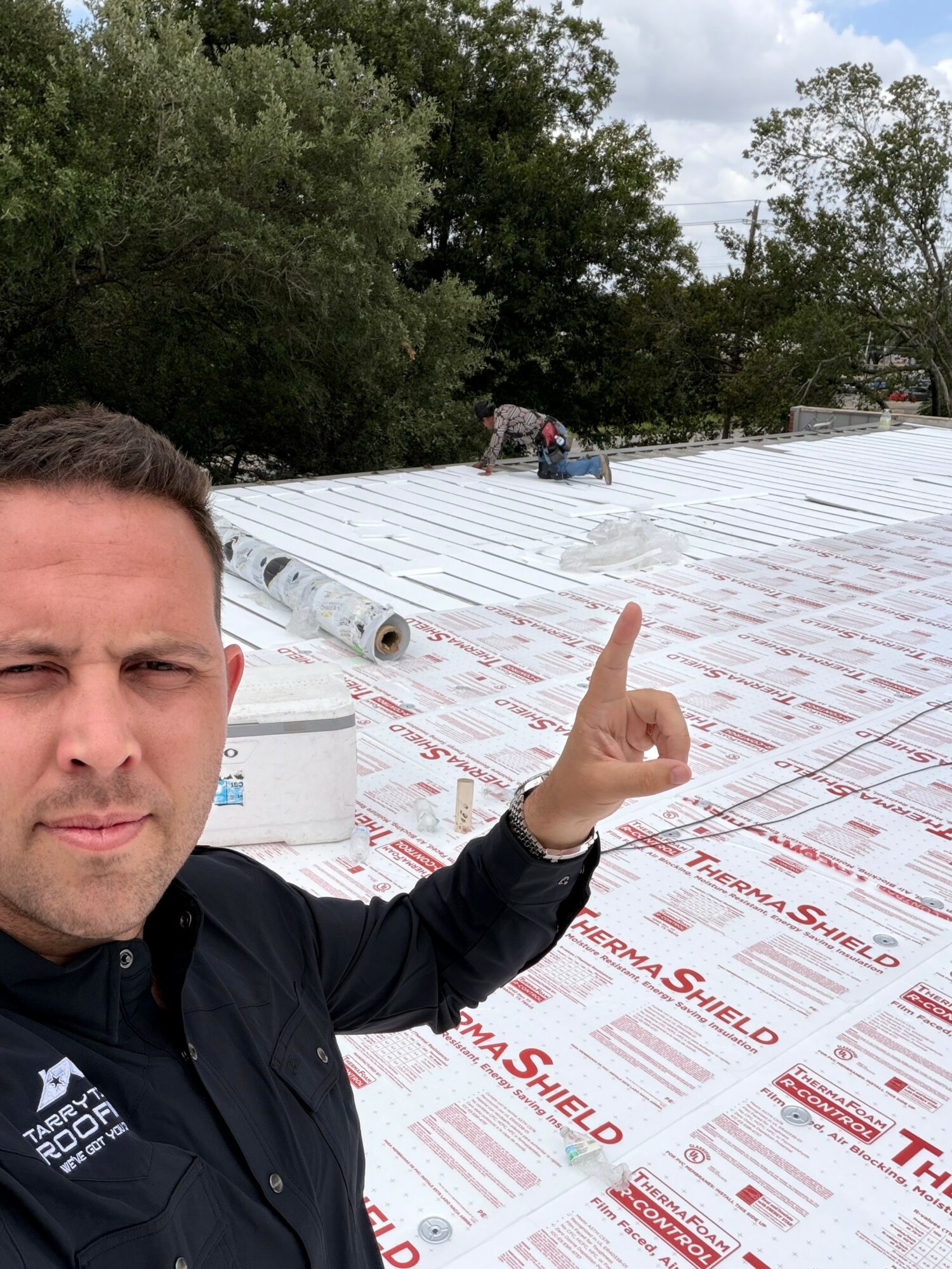 Man taking selfie outdoors on a roof with construction materials, pointing upward, trees in background, cloudy sky.