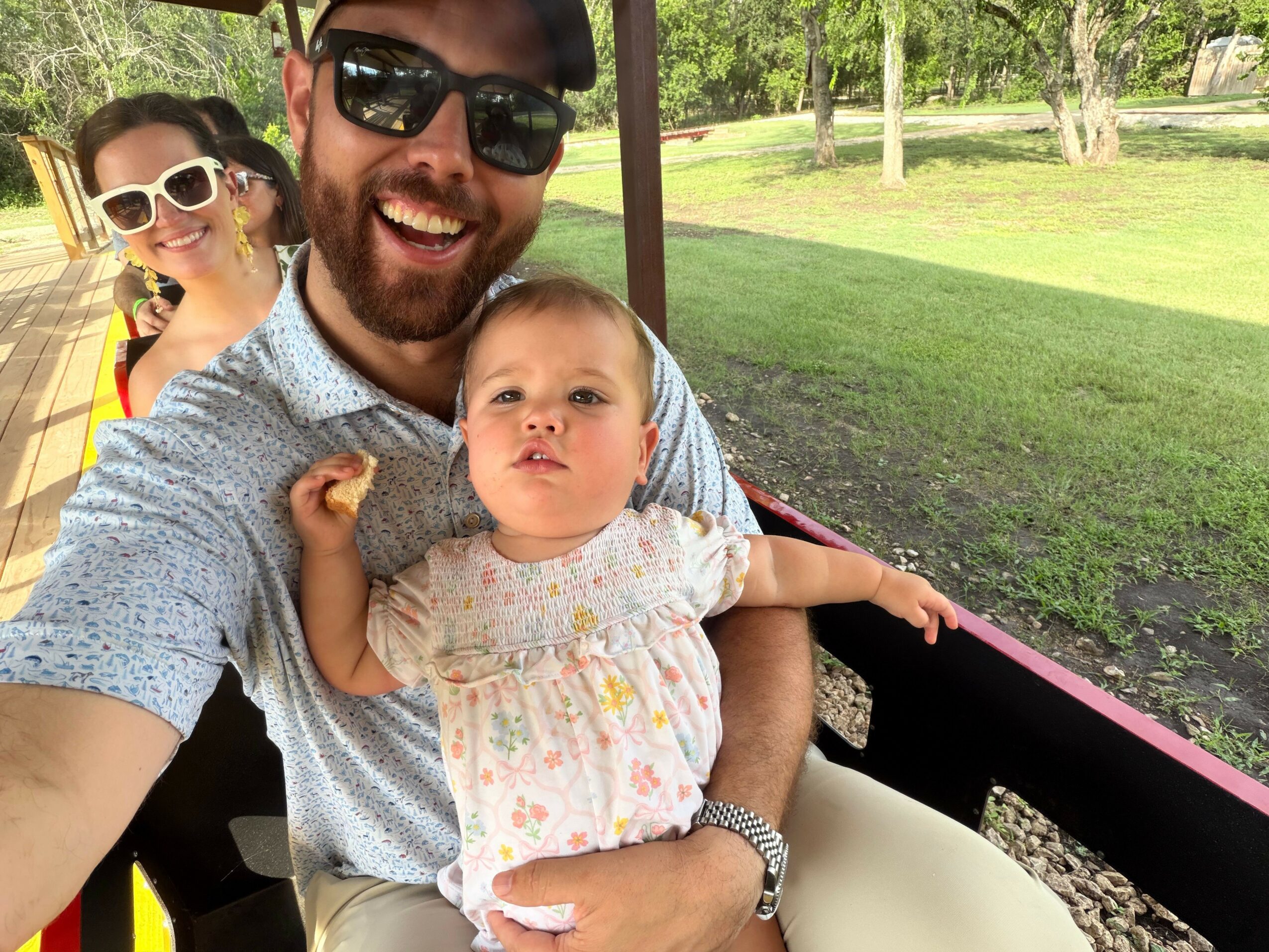 Man with sunglasses and a child with sunglasses smiling outdoors, with a young girl leaning on his shoulder, sitting at a table.